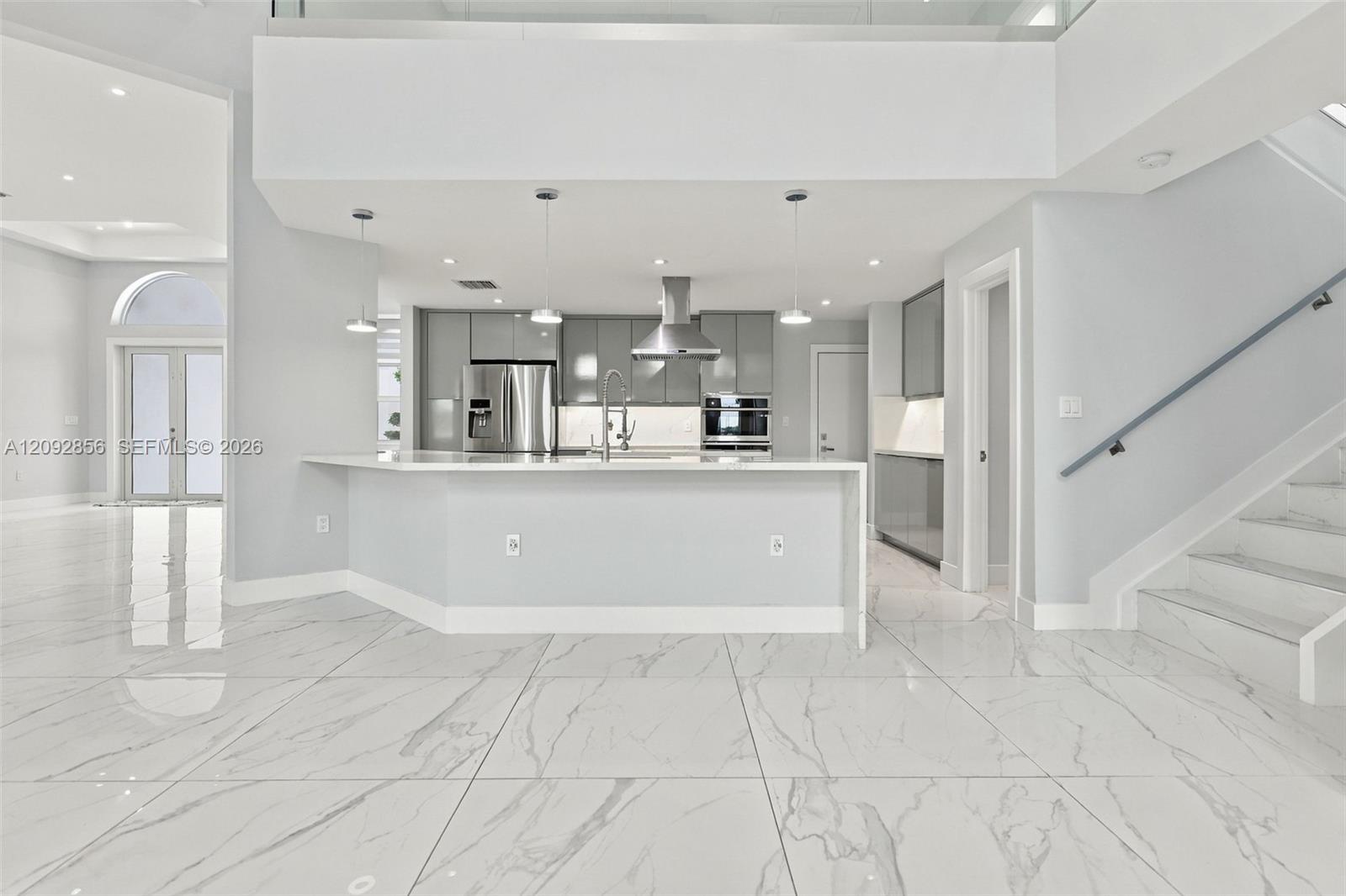 Open-concept kitchen featuring large-format tile flooring, gray gloss cabinetry, a stainless steel range hood, quartz countertops, and a glass-paneled staircase railing