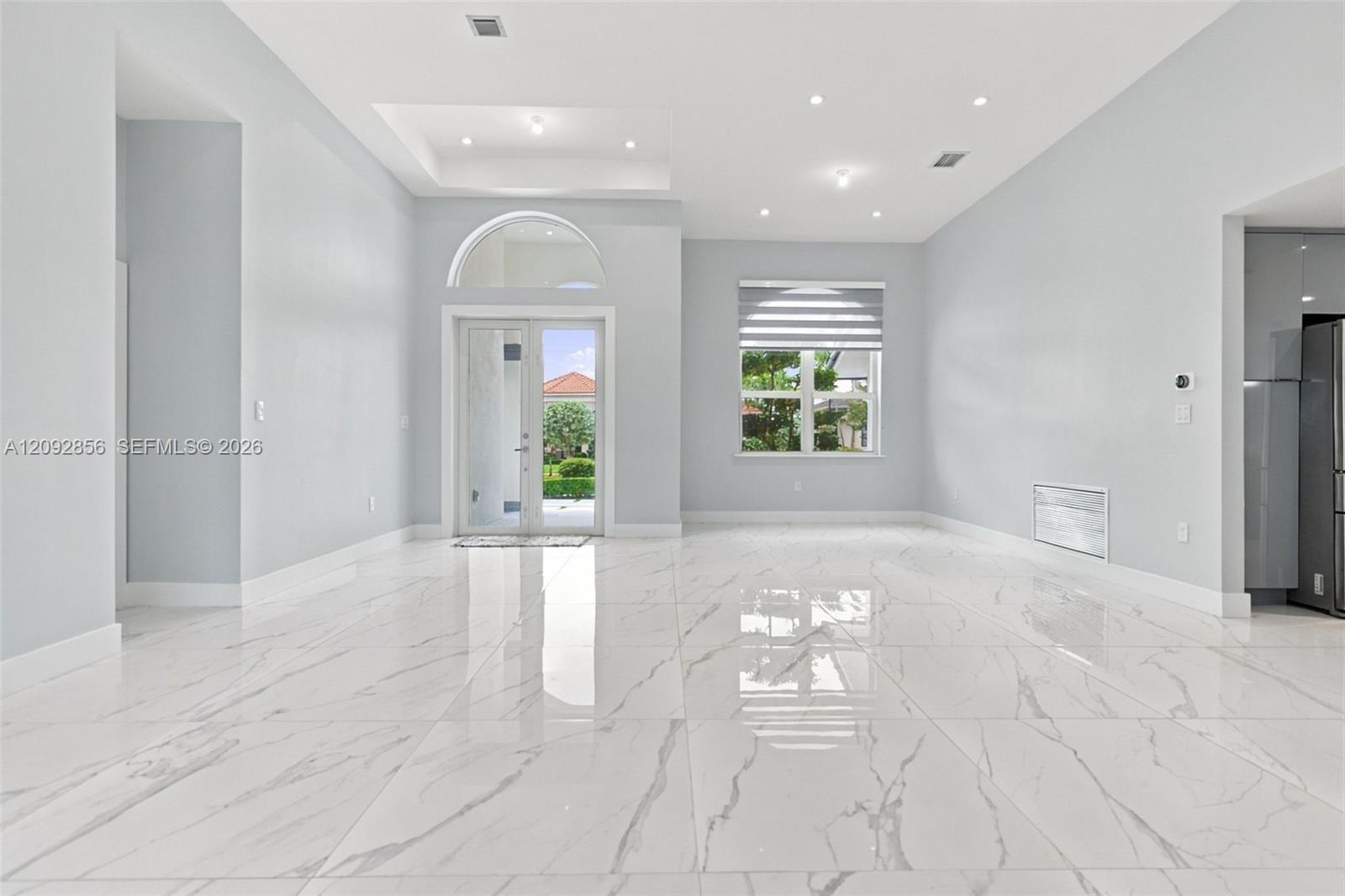 Open-concept interior featuring large-format polished stone-finish flooring, recessed lighting, a tray ceiling, and a glass-paneled double door with an arched transom