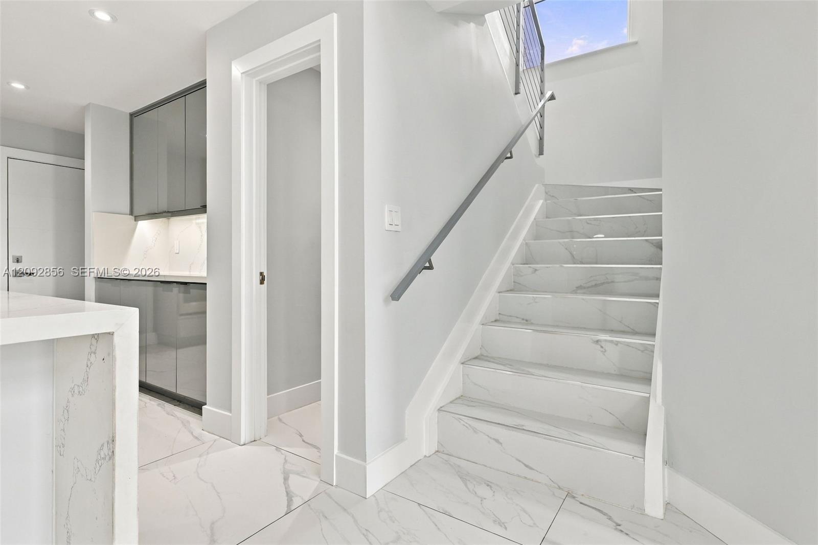 Staircase featuring stone-finish treads, a metal handrail, and white-finish wall accents, paired with nearby cabinetry featuring sleek hardware and integrated lighting