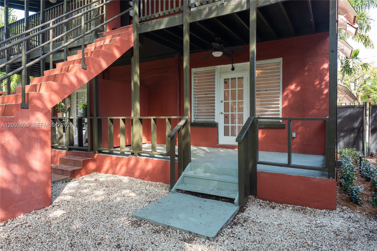 Exterior covered patio featuring stucco siding, a multi-pane glass door, dark-finished wooden railings, a ceiling fan, and concrete stairs
