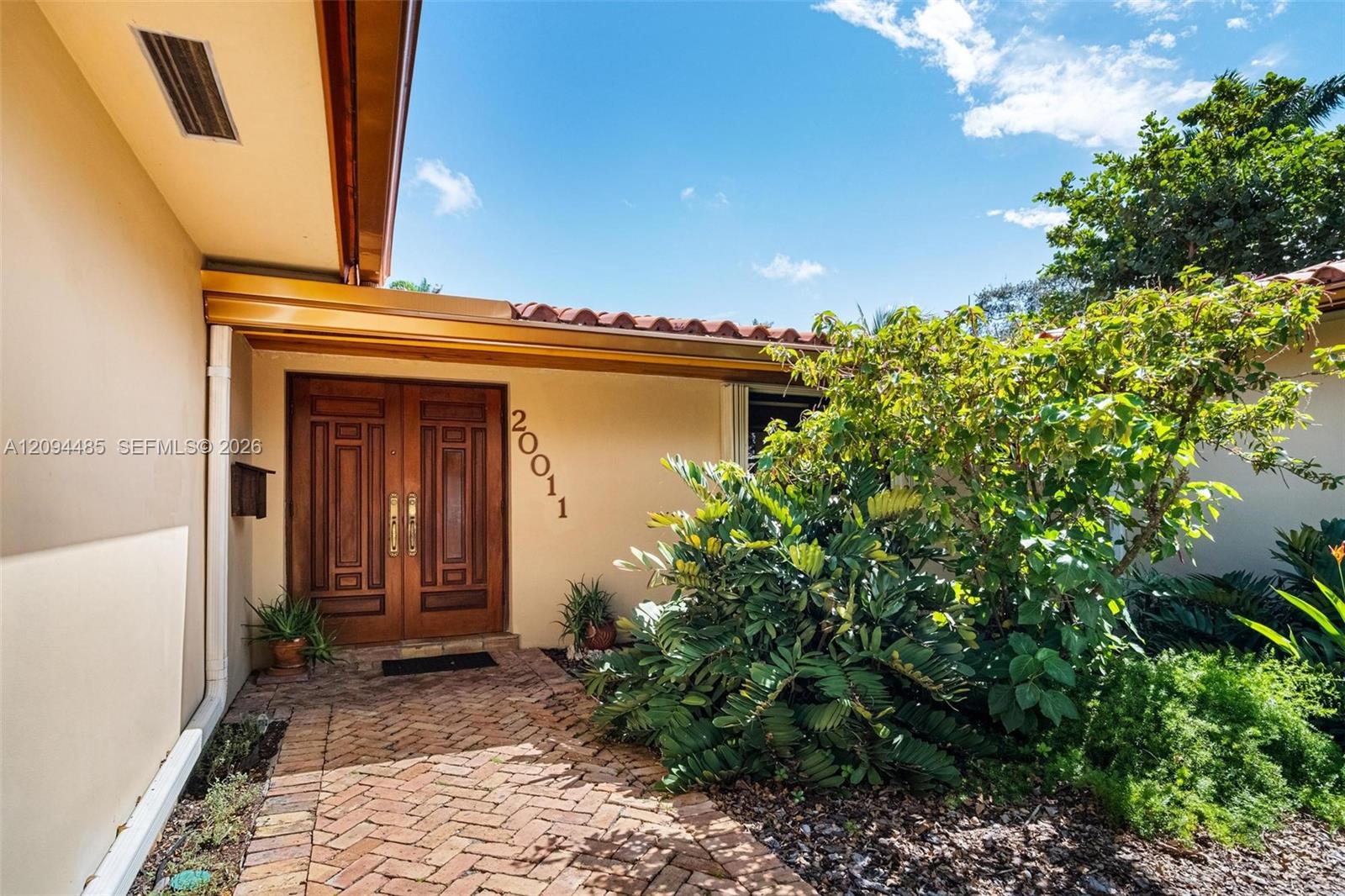 Stucco exterior entry featuring a herringbone-patterned brick walkway, double wood-paneled doors with metallic hardware, and a terracotta tile roof edge Stucco exterior entry featuring a herringbone-patterned brick walkway, double wood-paneled doors with metallic hardware, and a terracotta tile roof edge