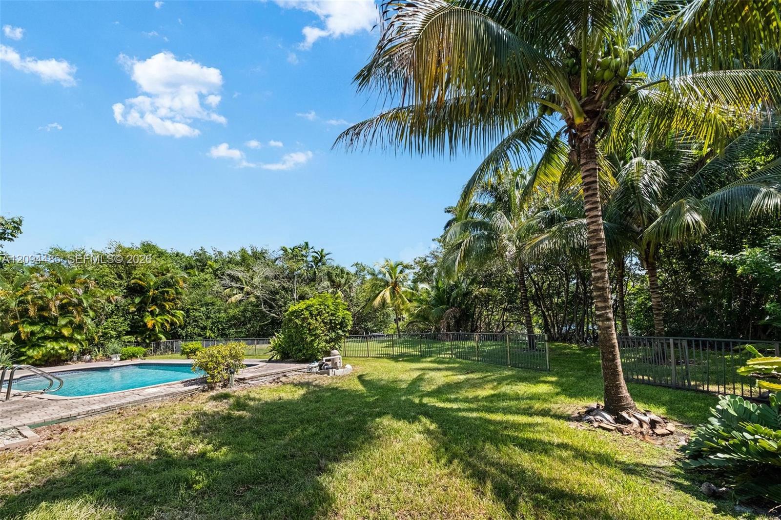 Backyard featuring an in-ground swimming pool, concrete pool deck, mature palm trees, perimeter fencing, and expansive green space Backyard featuring an in-ground swimming pool, concrete pool deck, mature palm trees, perimeter fencing, and expansive green space