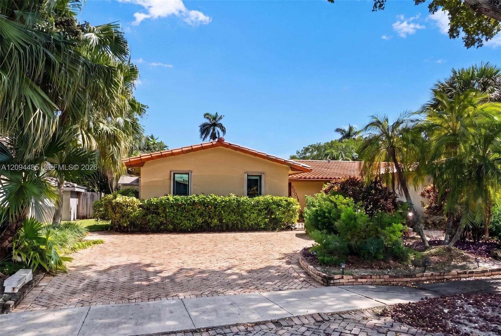 Single-story residence featuring a terracotta-tiled roof, brick-paved driveway, and lush tropical landscaping Single-story residence featuring a terracotta-tiled roof, brick-paved driveway, and lush tropical landscaping