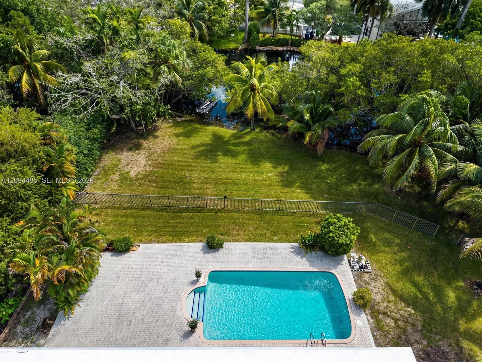 Outdoor living space featuring an in-ground swimming pool, concrete paver patio, metal perimeter fencing, manicured lawn, and mature tropical landscaping Outdoor living space featuring an in-ground swimming pool, concrete paver patio, metal perimeter fencing, manicured lawn, and mature tropical landscaping