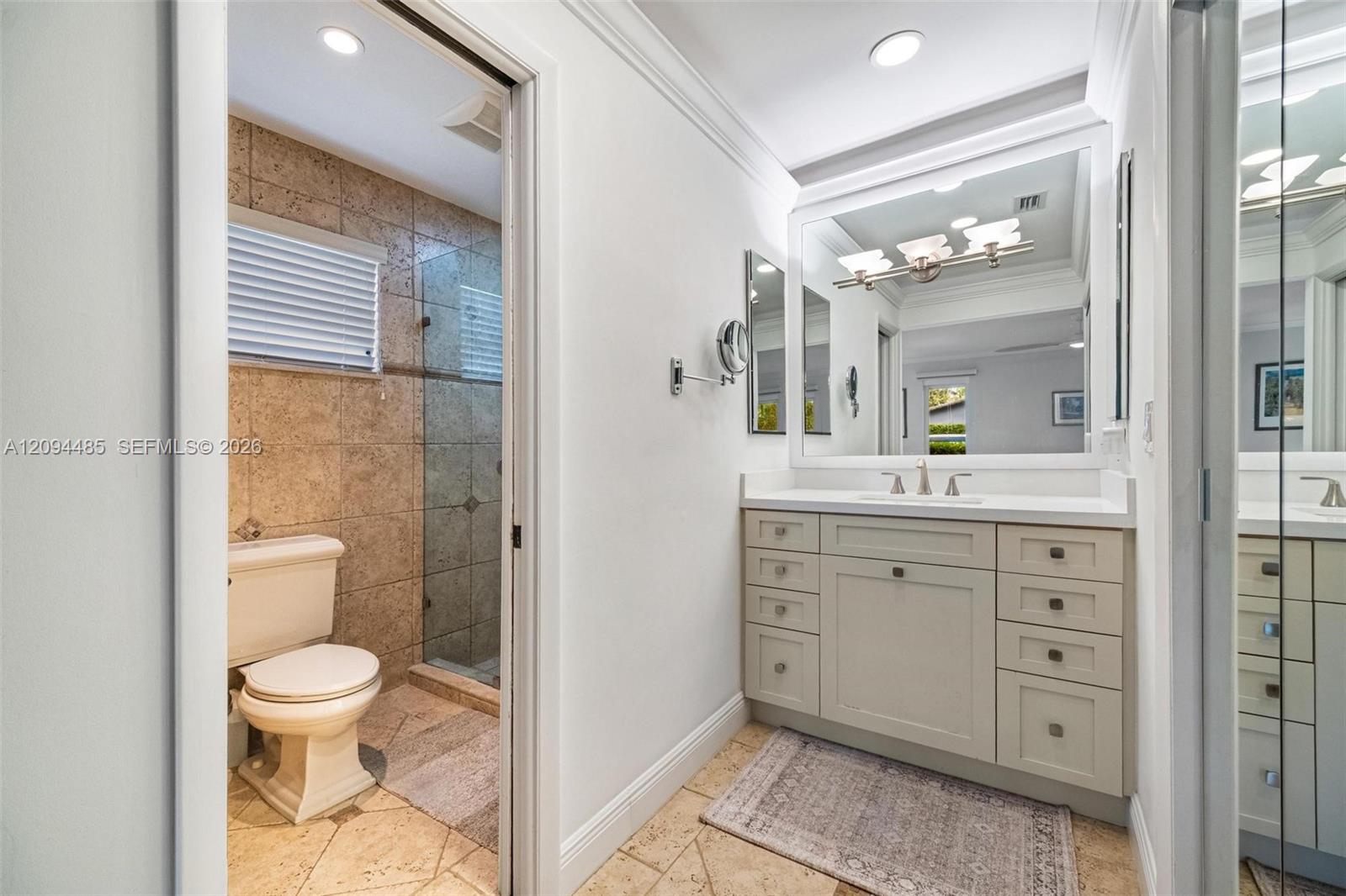 Vanity featuring a white countertop with an integrated basin, shaker-style cabinetry with metallic hardware, large-format wall mirror, and natural stone tile flooring