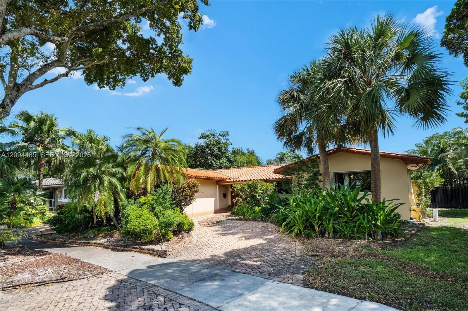 Single-story residence featuring a terracotta-tiled roof, stucco exterior, and a herringbone-patterned brick paver driveway, surrounded by established landscaping and mature palm trees Single-story residence featuring a terracotta-tiled roof, stucco exterior, and a herringbone-patterned brick paver driveway, surrounded by established landscaping and mature palm trees