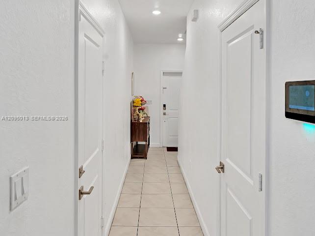 Hallway featuring neutral tile flooring, recessed ceiling lighting, wall-mounted smart home control panel, and solid-core interior doors with satin nickel lever handles Hallway featuring neutral tile flooring, recessed ceiling lighting, wall-mounted smart home control panel, and solid-core interior doors with satin nickel lever handles