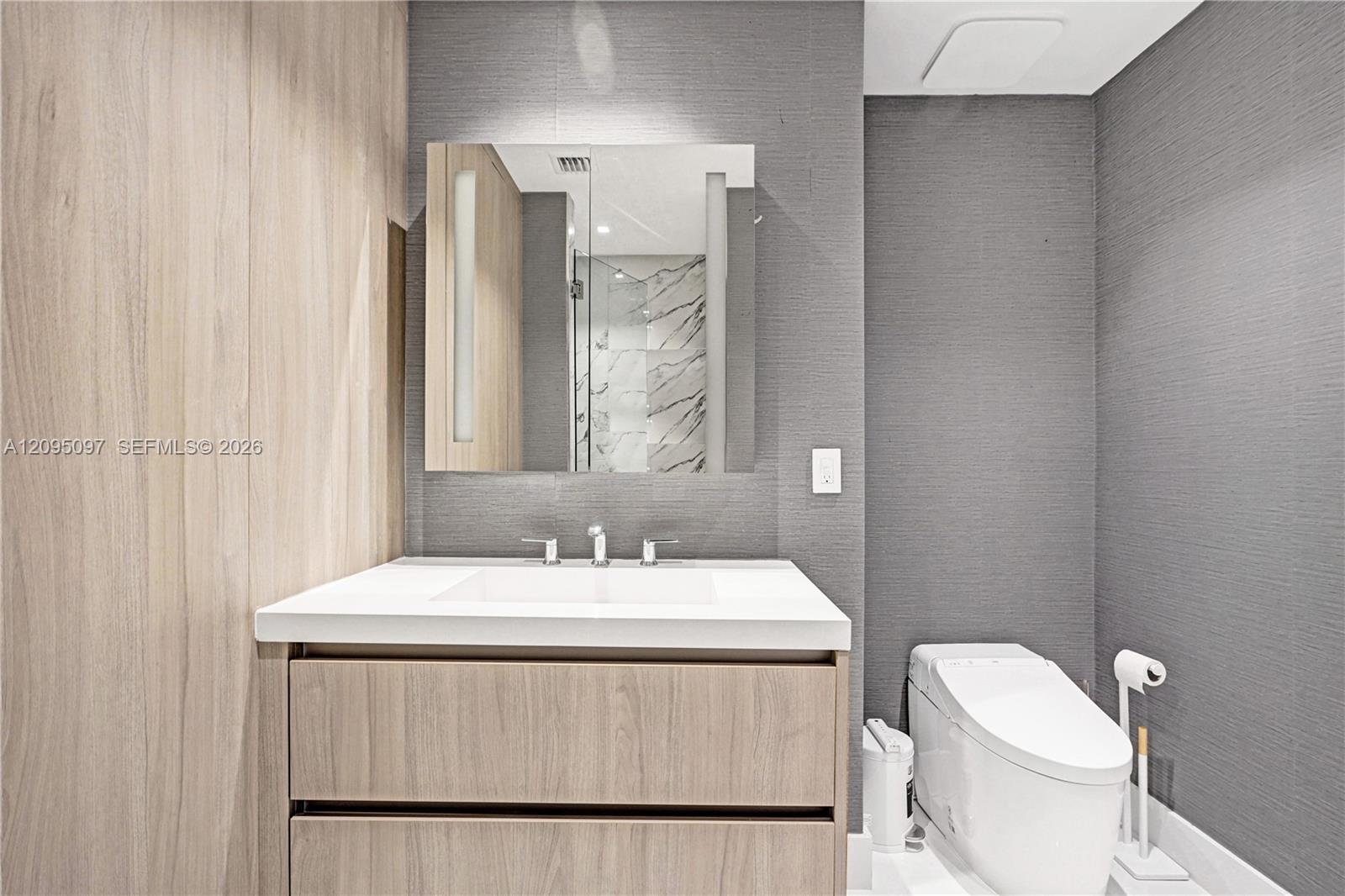 Floating vanity with wood-finish cabinetry, crisp white countertop, integrated rectangular sink, wall-mounted mirror with integrated lighting, and textured grey wall finishes