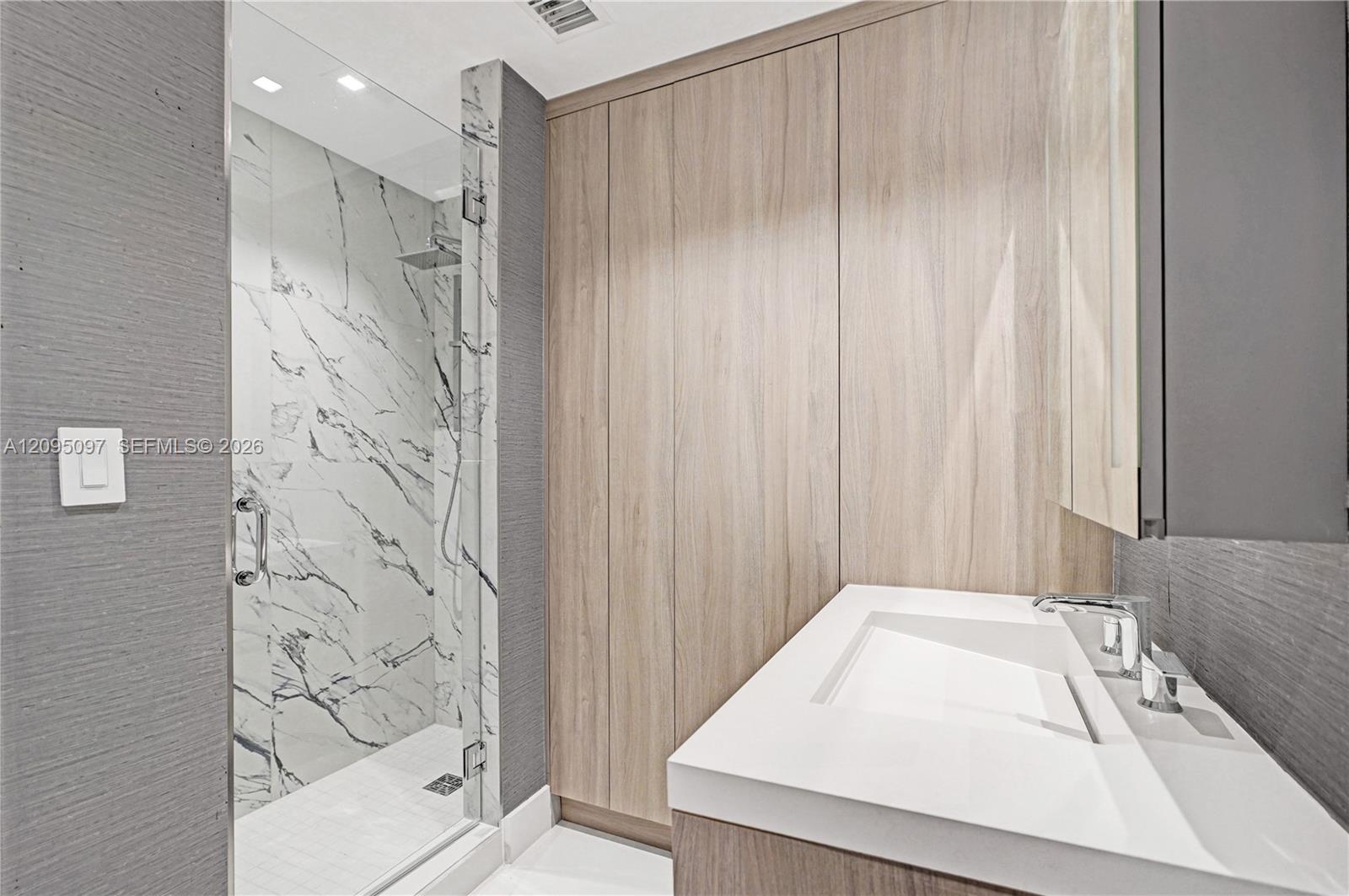 Modern bathroom featuring textured wall surfaces, integrated wood-finish cabinetry, a glass-enclosed shower with large-format veined tile, and a built-in vanity with a sleek solid-surface countertop and recessed basin
