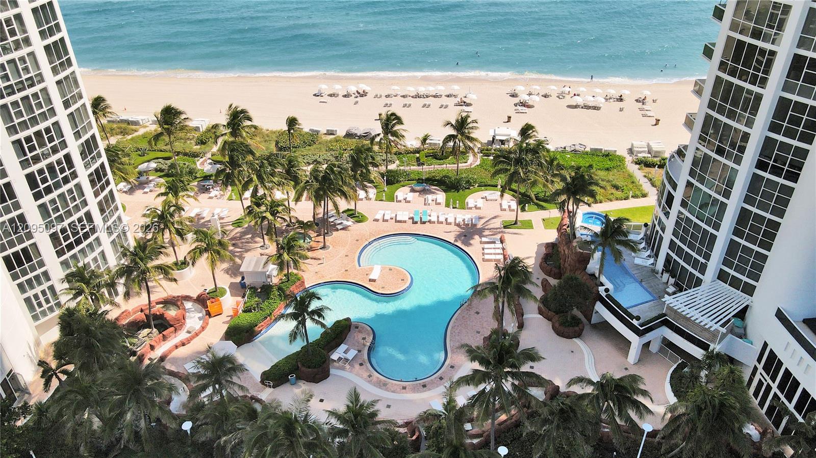 Aerial perspective of property grounds featuring a free-form swimming pool, integrated hot tub, paved sun deck, mature palm trees, and coastal access
