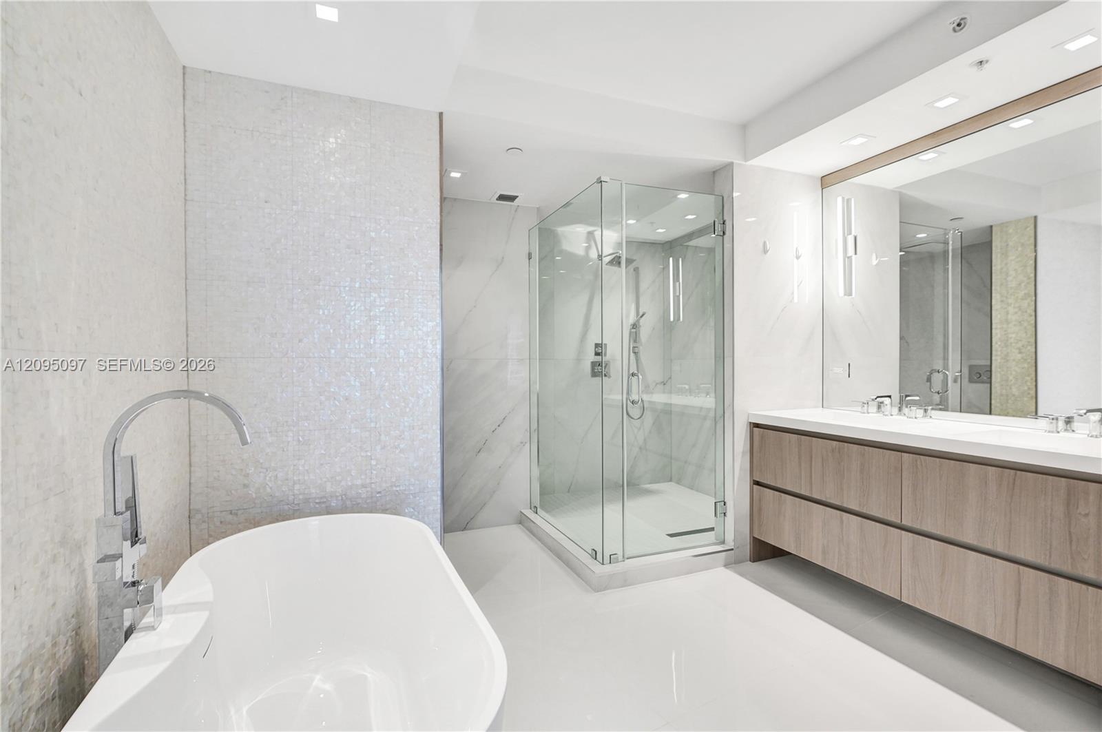 Freestanding soaking tub with floor-mounted faucet, glass-enclosed shower, wall-to-wall vanity with double sinks, and recessed lighting