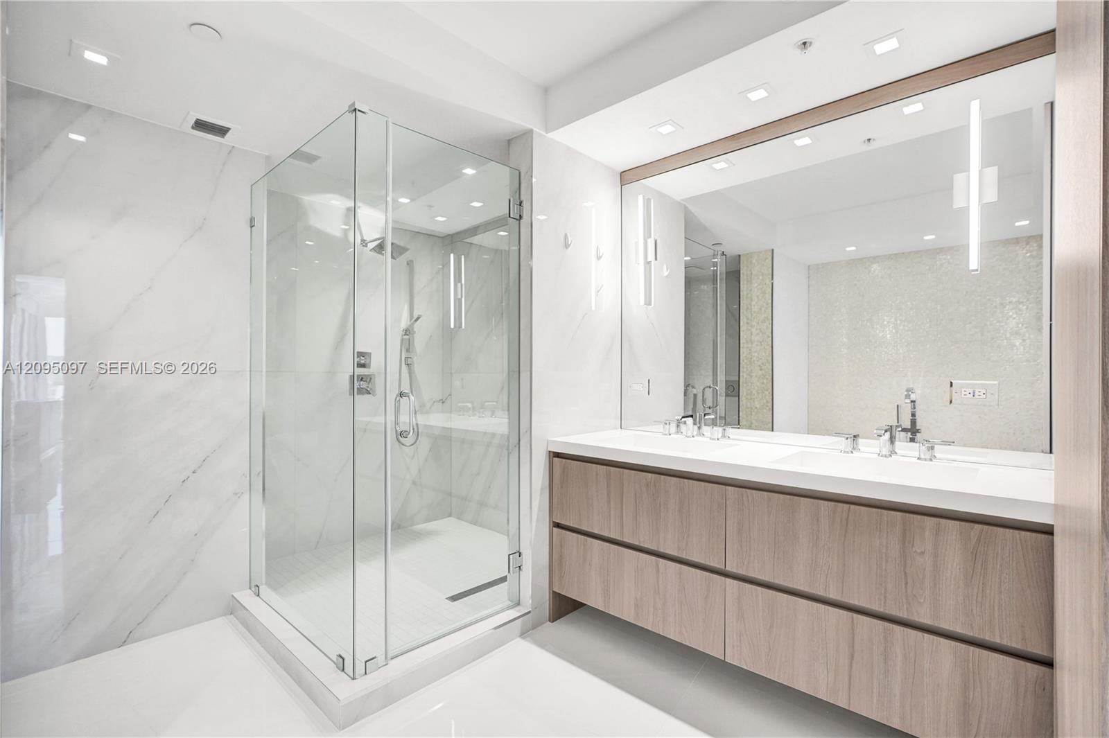 Floating vanity with dual rectangular sinks, frameless glass-enclosed shower, large-format wall tiling, recessed lighting, and integrated wall mirror