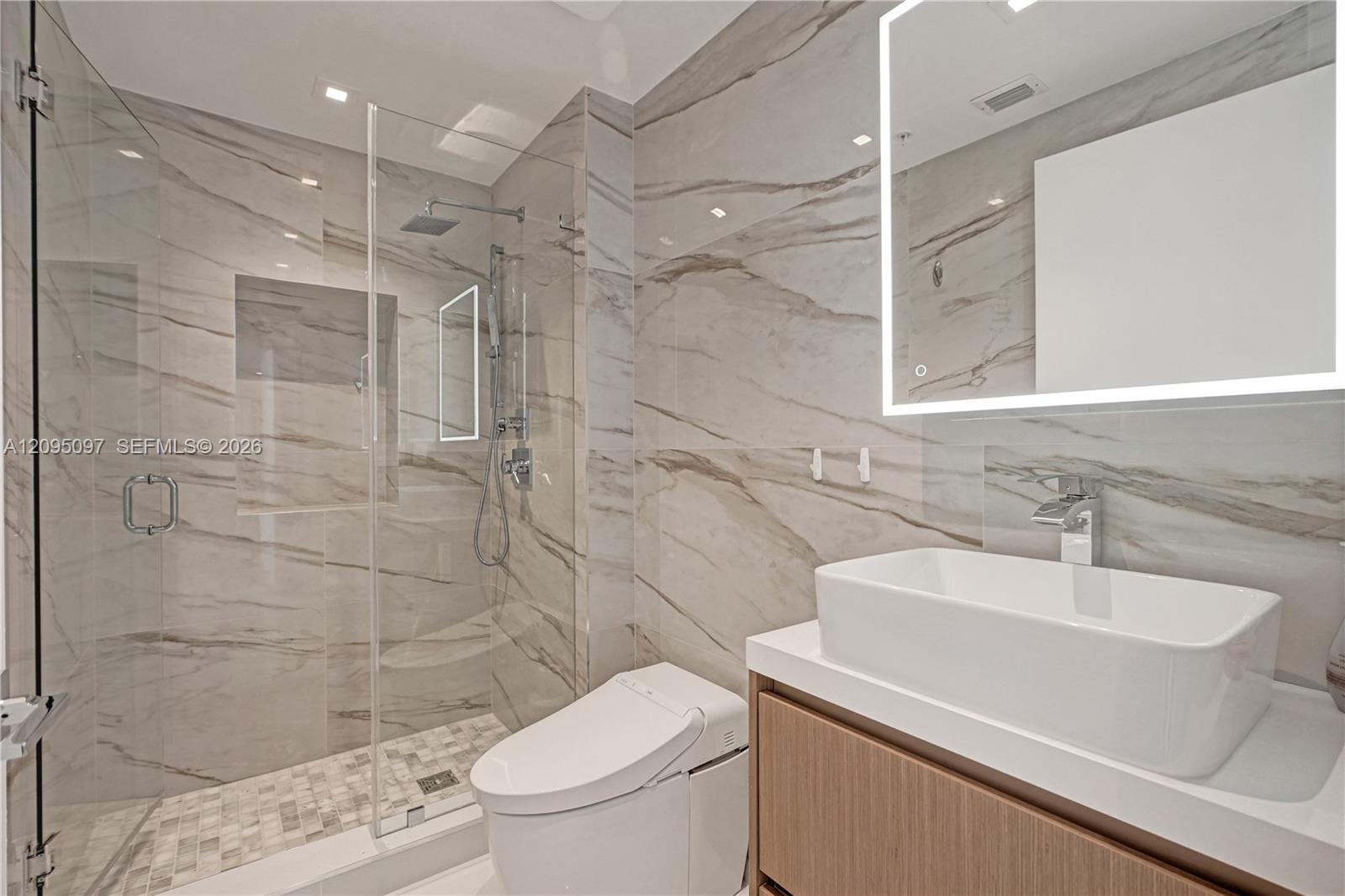 Bathroom featuring large-format wall tile, glass-enclosed shower with rainfall showerhead, built-in shower niche, floating wood-finish vanity with vessel sink, and illuminated wall-mounted mirror