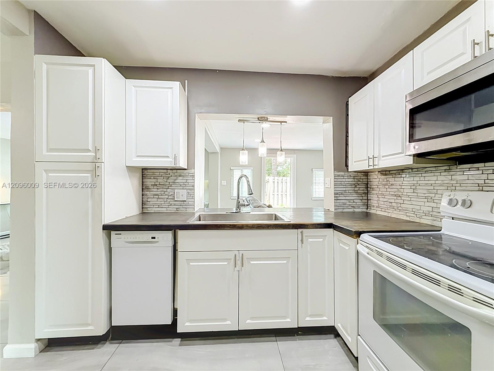 Kitchen featuring white cabinetry, dark countertops, a stacked-stone tile backsplash, stainless steel sink with pull-down faucet, and white appliances