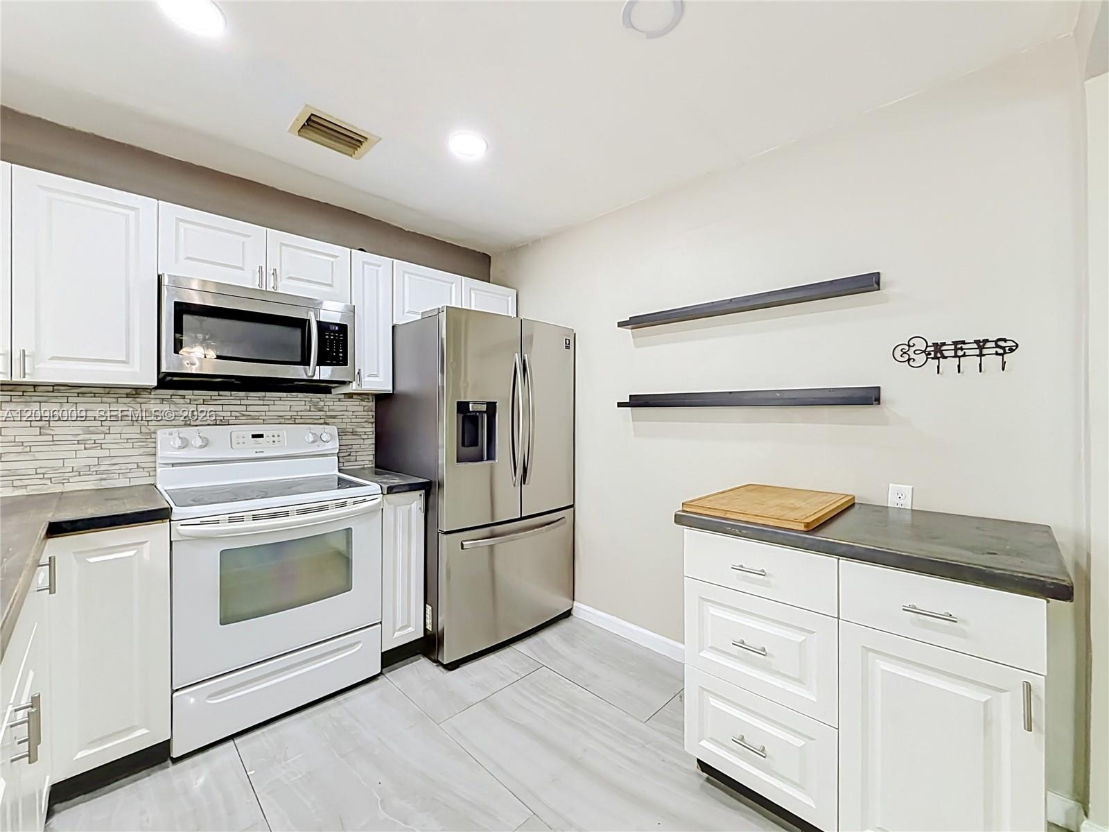 Kitchen featuring white cabinetry, stainless steel appliances, linear tile backsplash, dark solid-surface countertops, and light-toned tile flooring