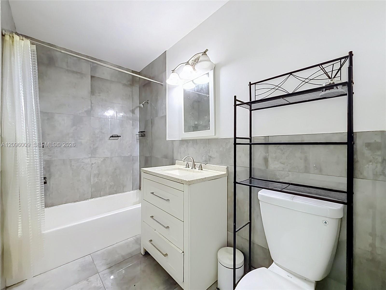 Bathroom featuring large-format grey tile surround, white vanity cabinet with stone-finish countertop, undermount sink, and three-light wall fixture