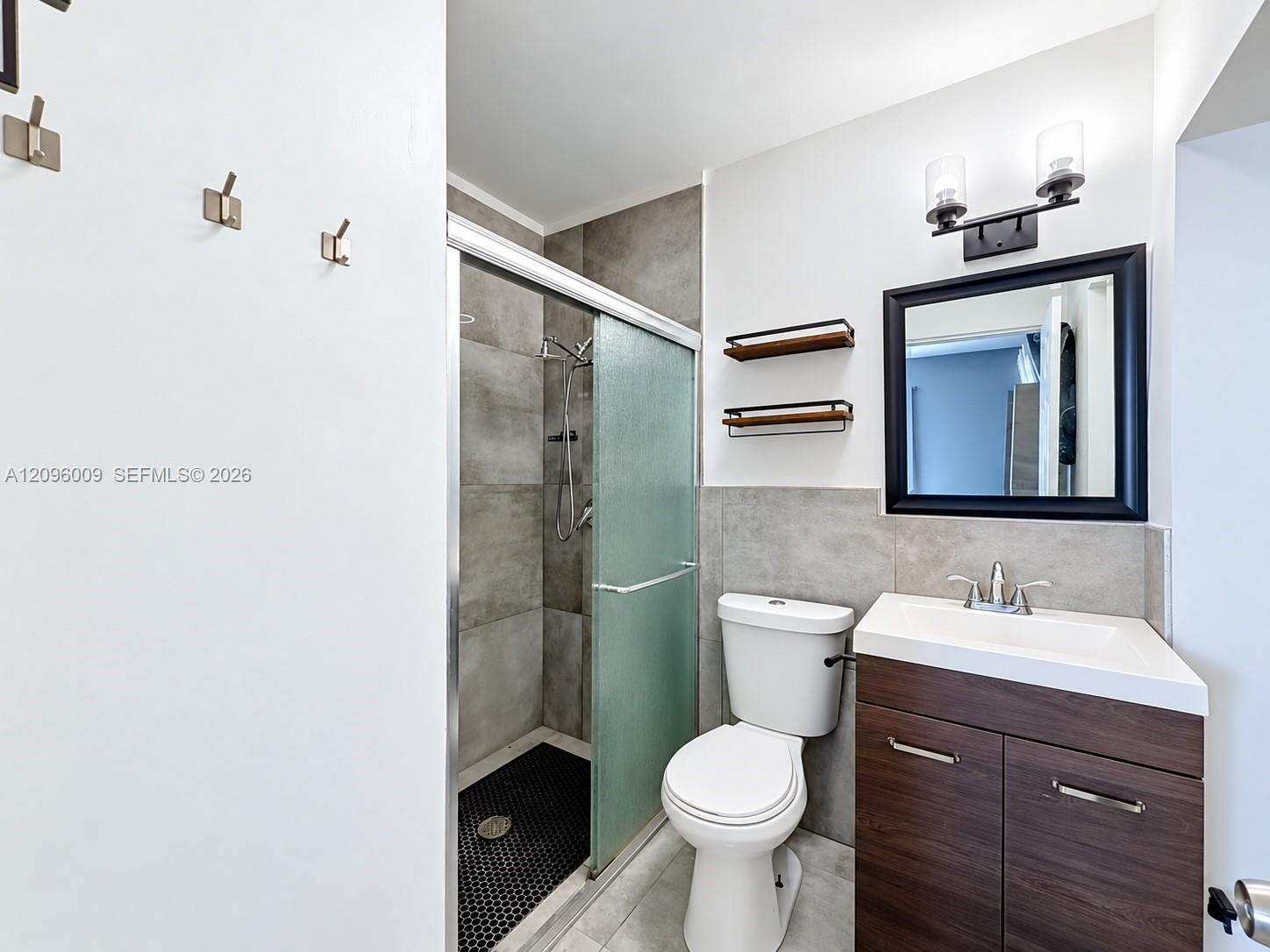 Bathroom featuring a walk-in shower with large-format tile surround, dark wood-finish vanity with integrated sink, two-light vanity fixture, and black-framed wall mirror