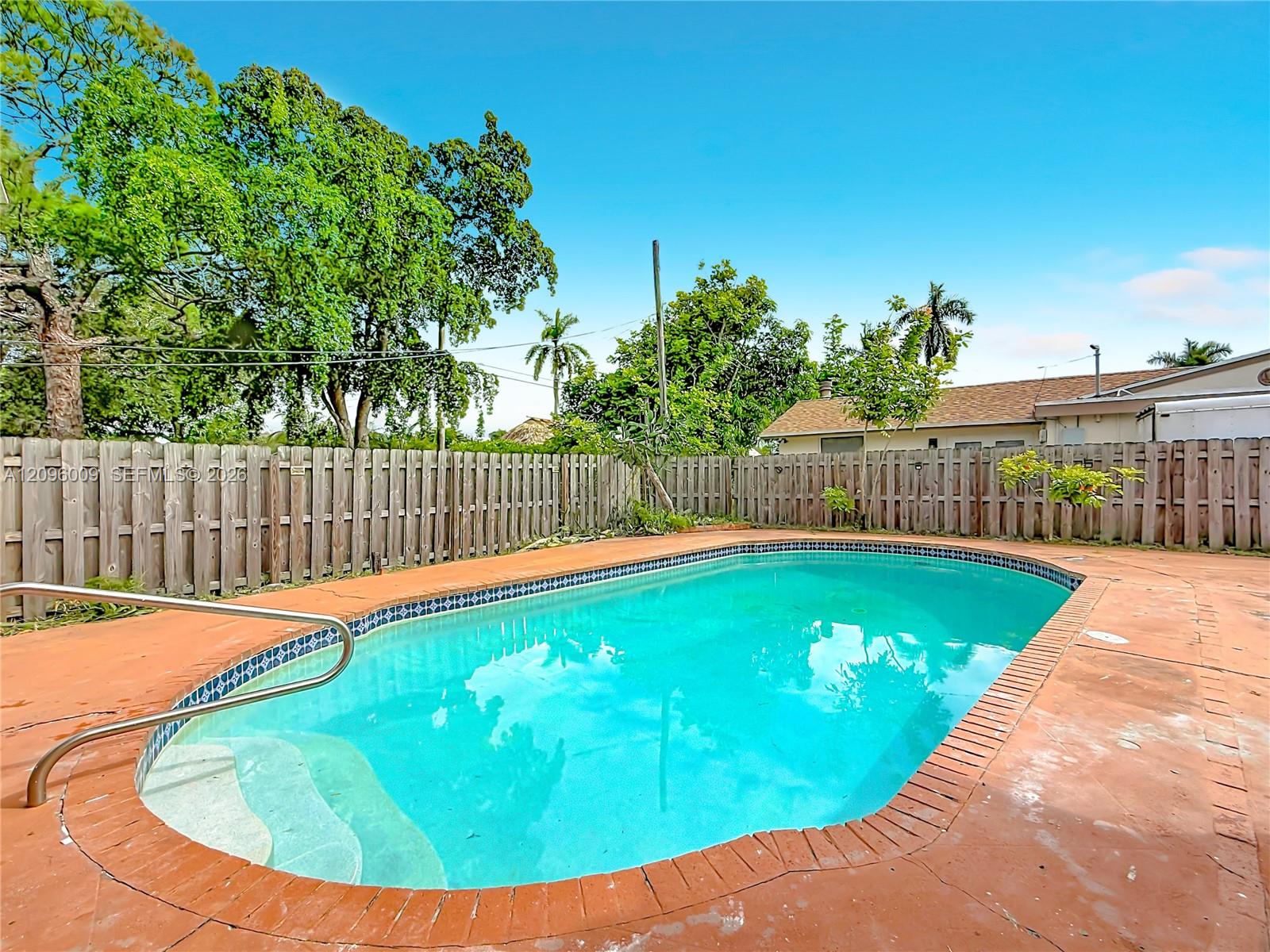 In-ground swimming pool featuring built-in entry steps, a metal handrail, brick-style coping, and a concrete deck enclosed by a wood privacy fence