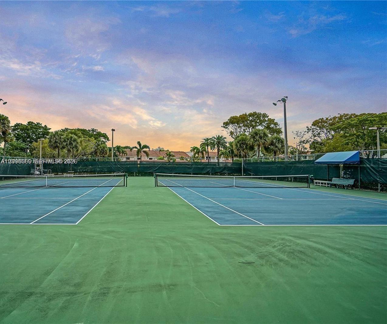 Two full-size tennis courts featuring hard-court surfaces, perimeter fencing, overhead lighting, and shaded bench seating