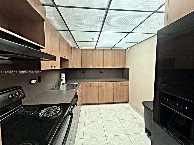 Kitchen featuring wood-finish cabinetry, neutral solid-surface countertops, a stainless steel undermount sink, integrated black appliances, and a grid-panel drop ceiling