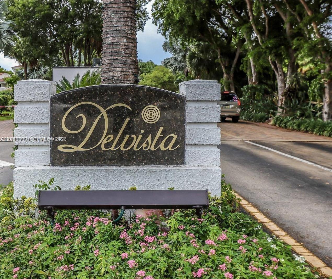 Community signage featuring a dark stone plaque with gold lettering, set within a textured white masonry monument base, surrounded by manicured landscaping and mature trees