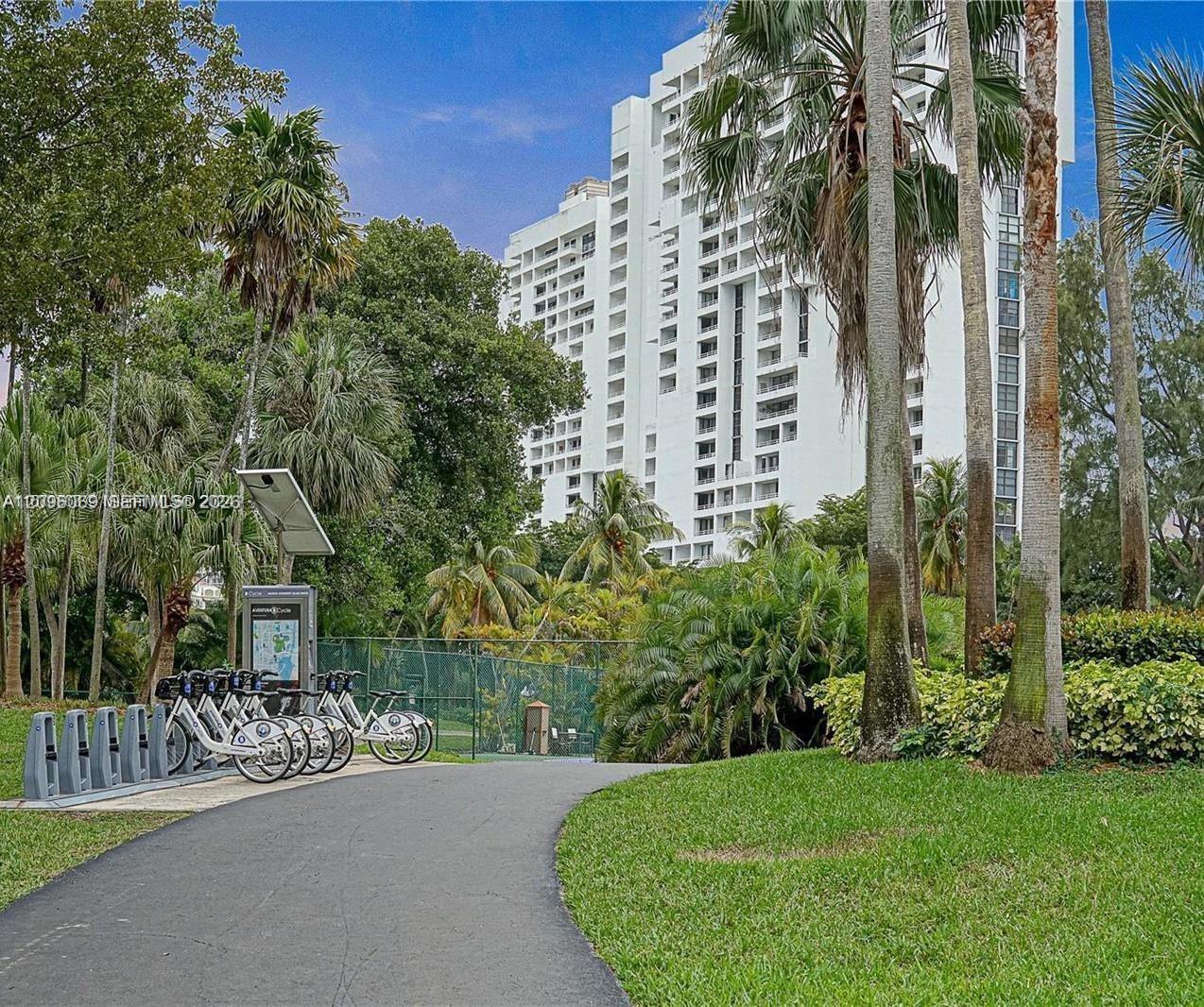High-rise residential exterior featuring a multi-story white facade with recessed balconies, paved pathways, mature palm trees, landscaped greenery, and a bicycle-sharing station