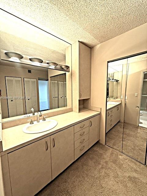 Bathroom featuring a vanity with integrated cabinetry, a recessed oval sink, a wall-to-wall mirror, overhead vanity lighting, and floor-to-ceiling mirrored closet doors