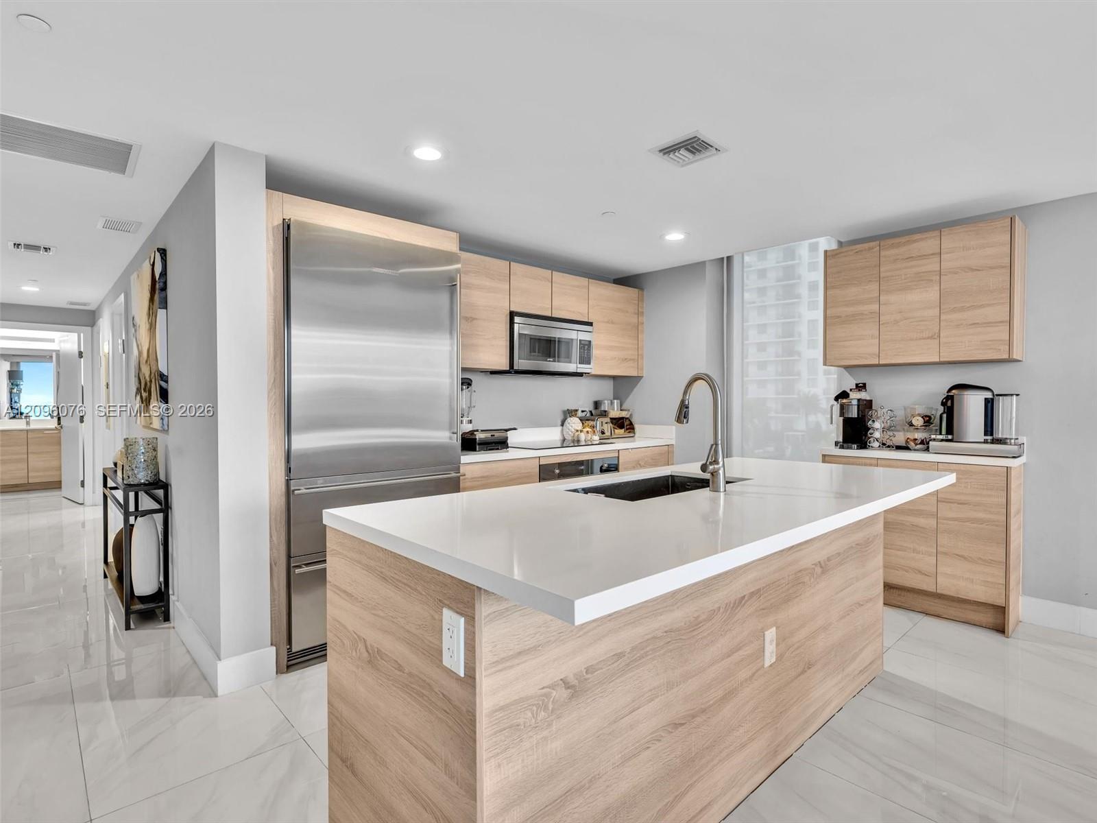Kitchen featuring a central island with a quartz countertop and under-mount sink, light-tone wood-finish cabinetry, stainless steel refrigerator, and large-format tile flooring