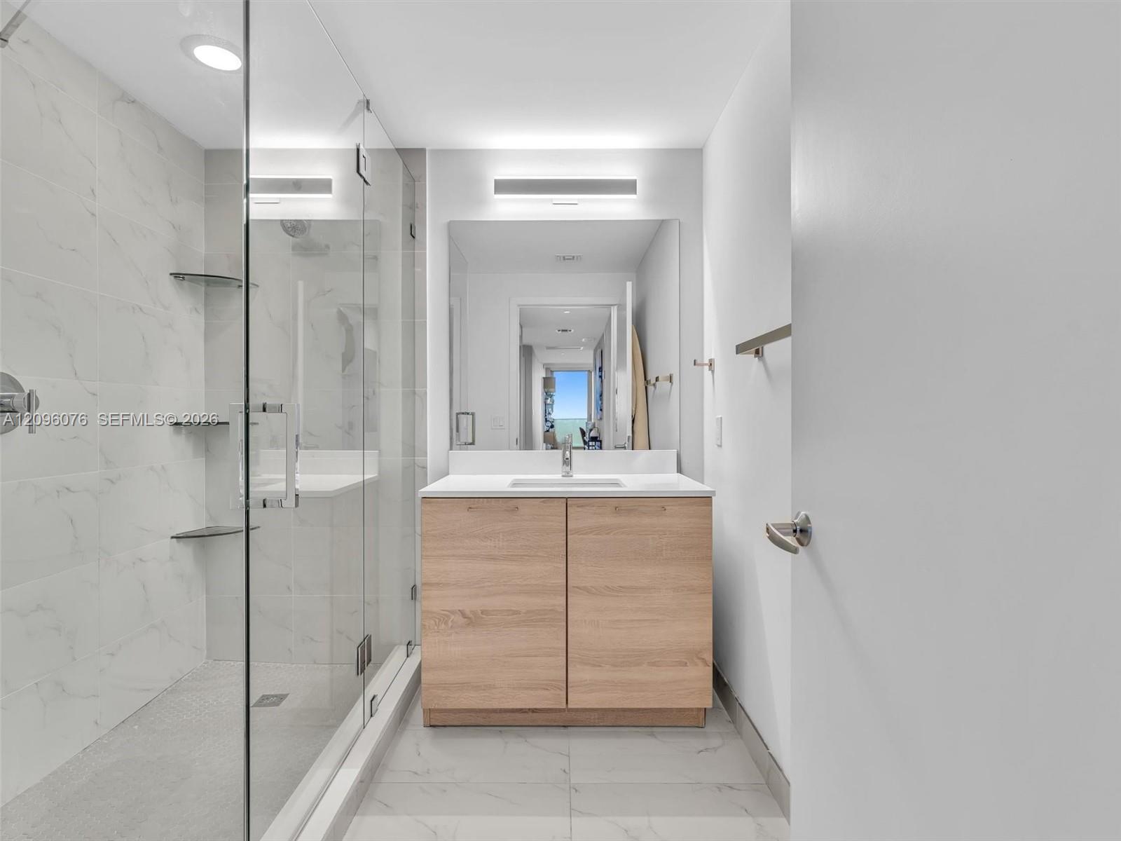 Walk-in shower with frameless glass enclosure, wood-finish cabinetry, white countertop with integrated sink, and large-format tile flooring