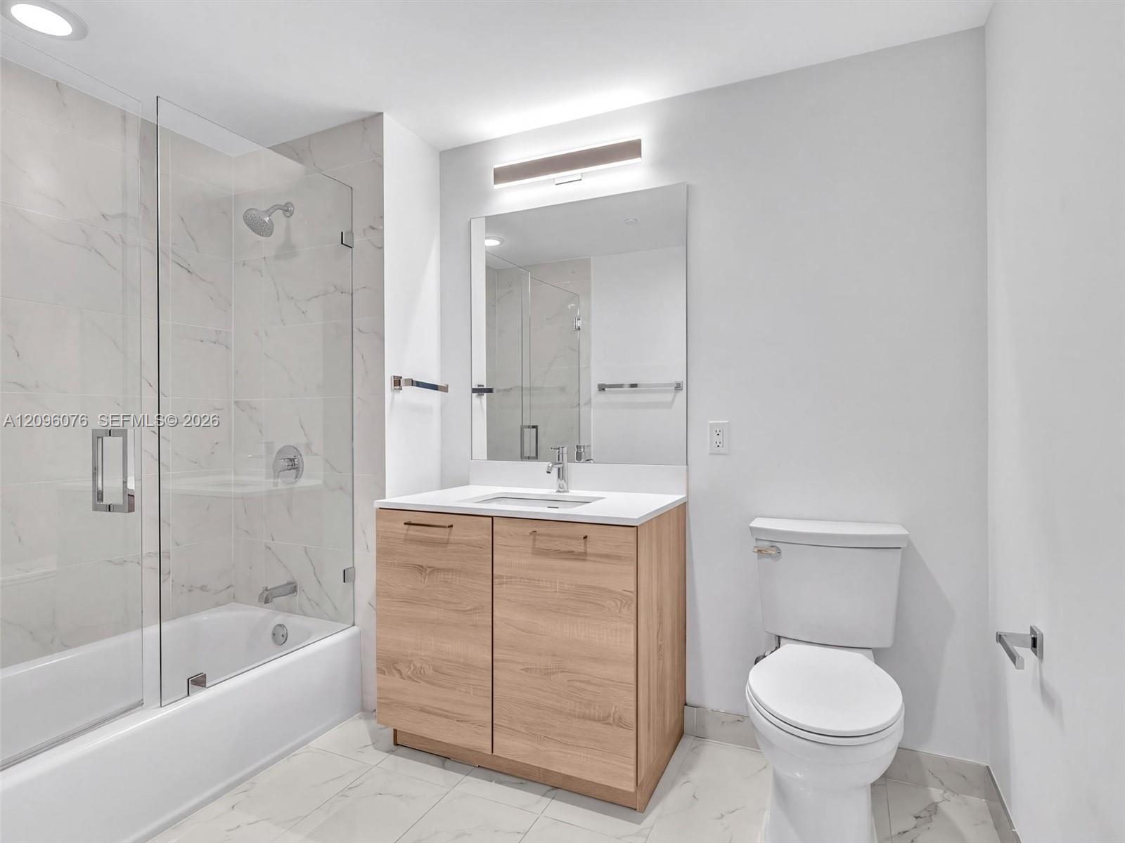 Bathroom featuring a wood-finish vanity with a stone countertop, a glass-enclosed shower and tub combination with tile surround, and large-format tile flooring