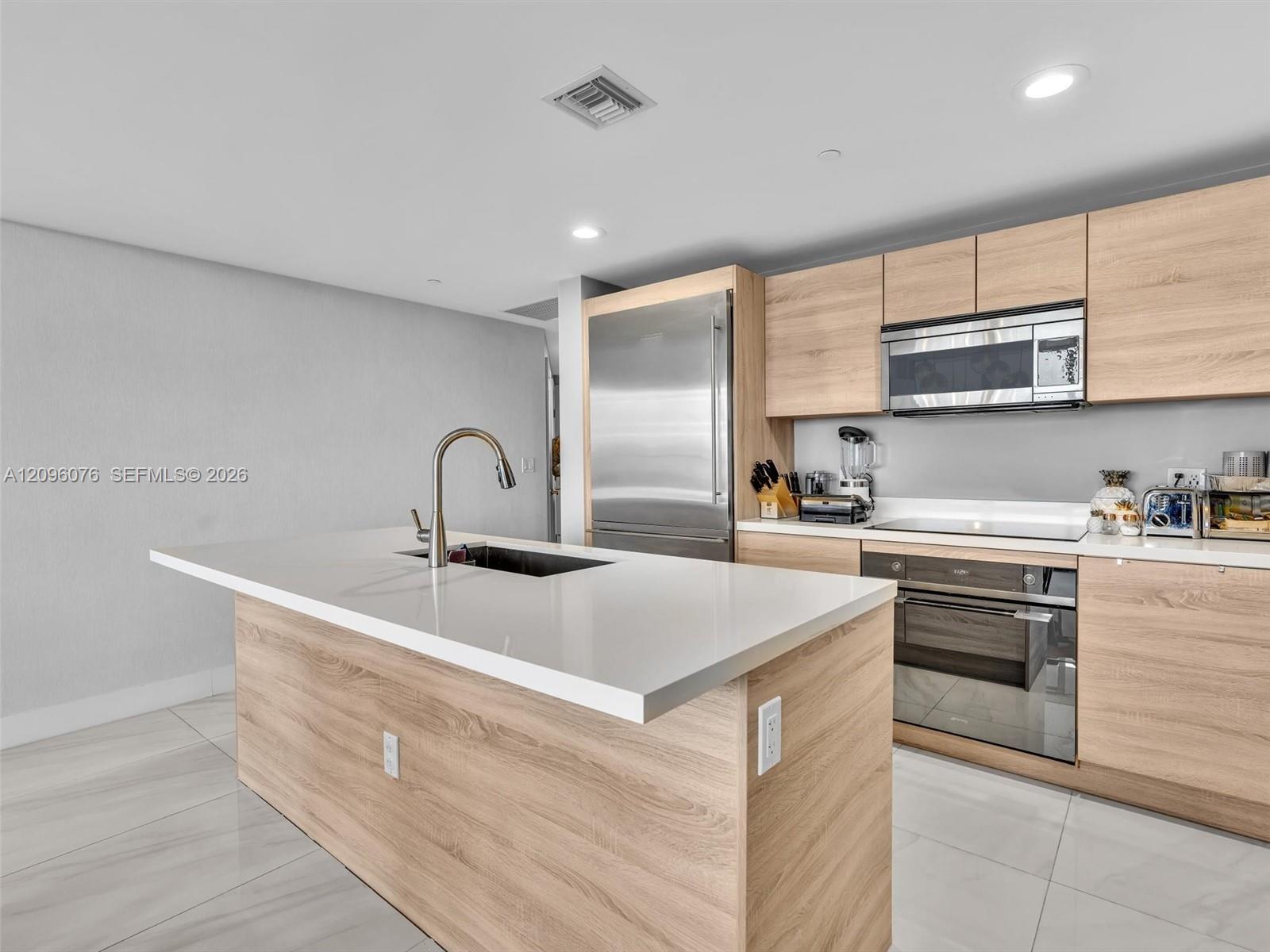 Kitchen featuring flat-panel wood-finish cabinetry, white composite countertops, a center island with an undermount sink, stainless steel appliances, and large-format tile flooring