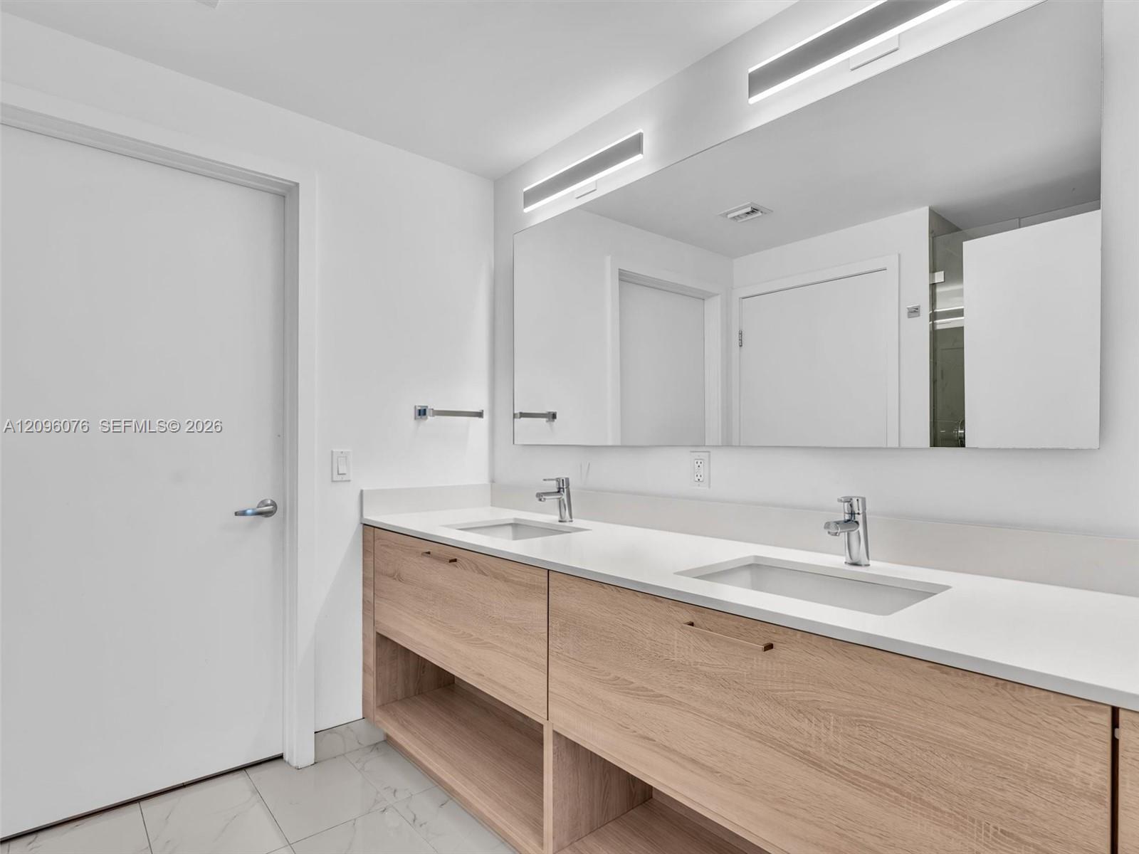 Dual-sink vanity featuring a light wood-finish cabinet with open shelving, integrated solid-surface countertop, rectangular undermount basins, and large wall-mounted mirror with recessed integrated lighting