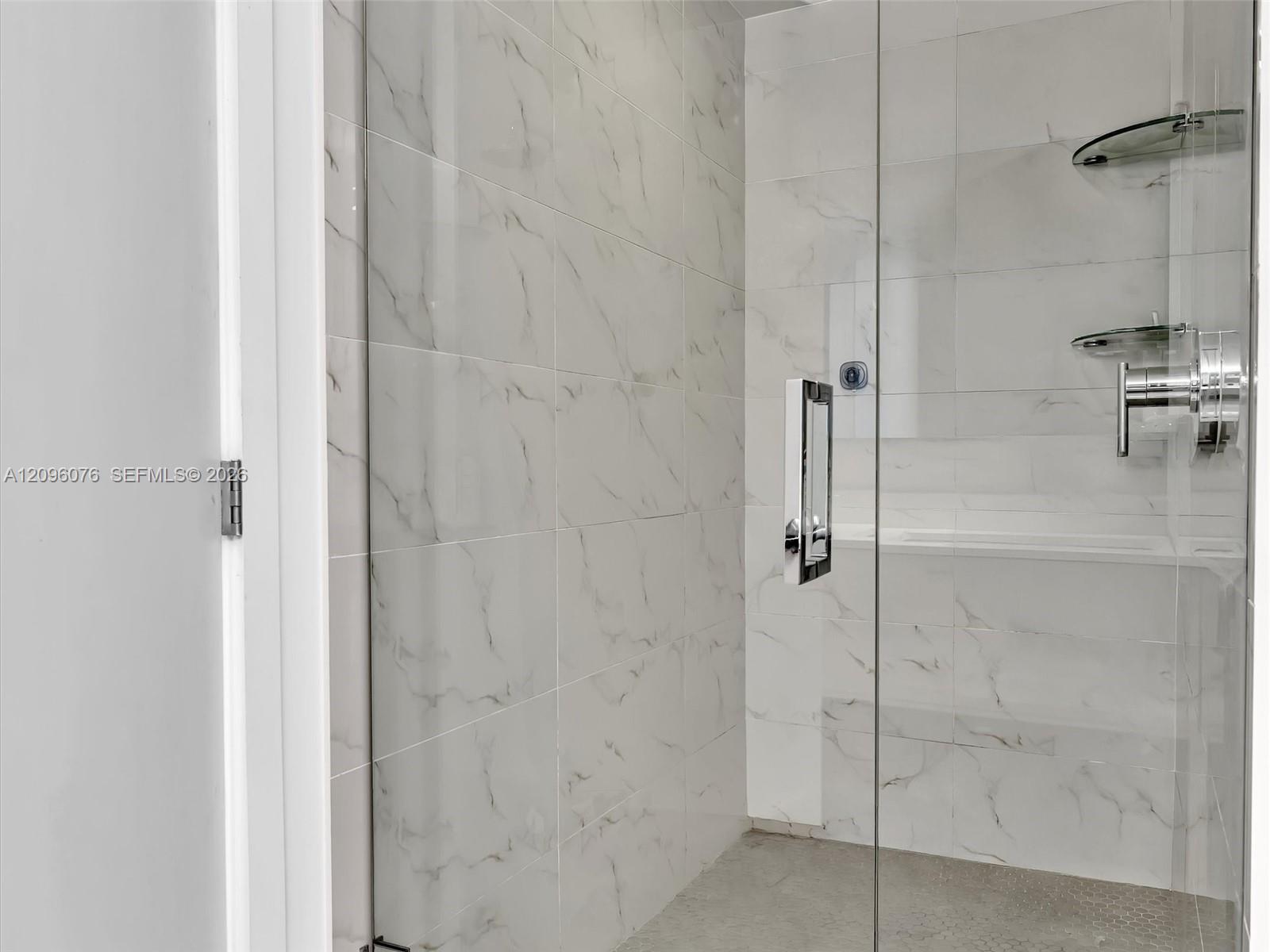 Walk-in shower featuring floor-to-ceiling veined tile, a frameless glass door, integrated glass shelving, and a polished metal shower fixture
