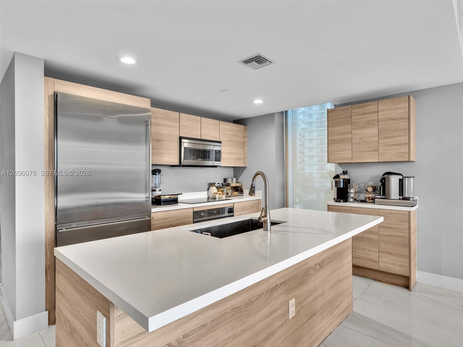 Kitchen featuring wood-finish cabinetry, quartz countertops, a central island with an integrated sink, stainless steel appliances, and large-format tile flooring