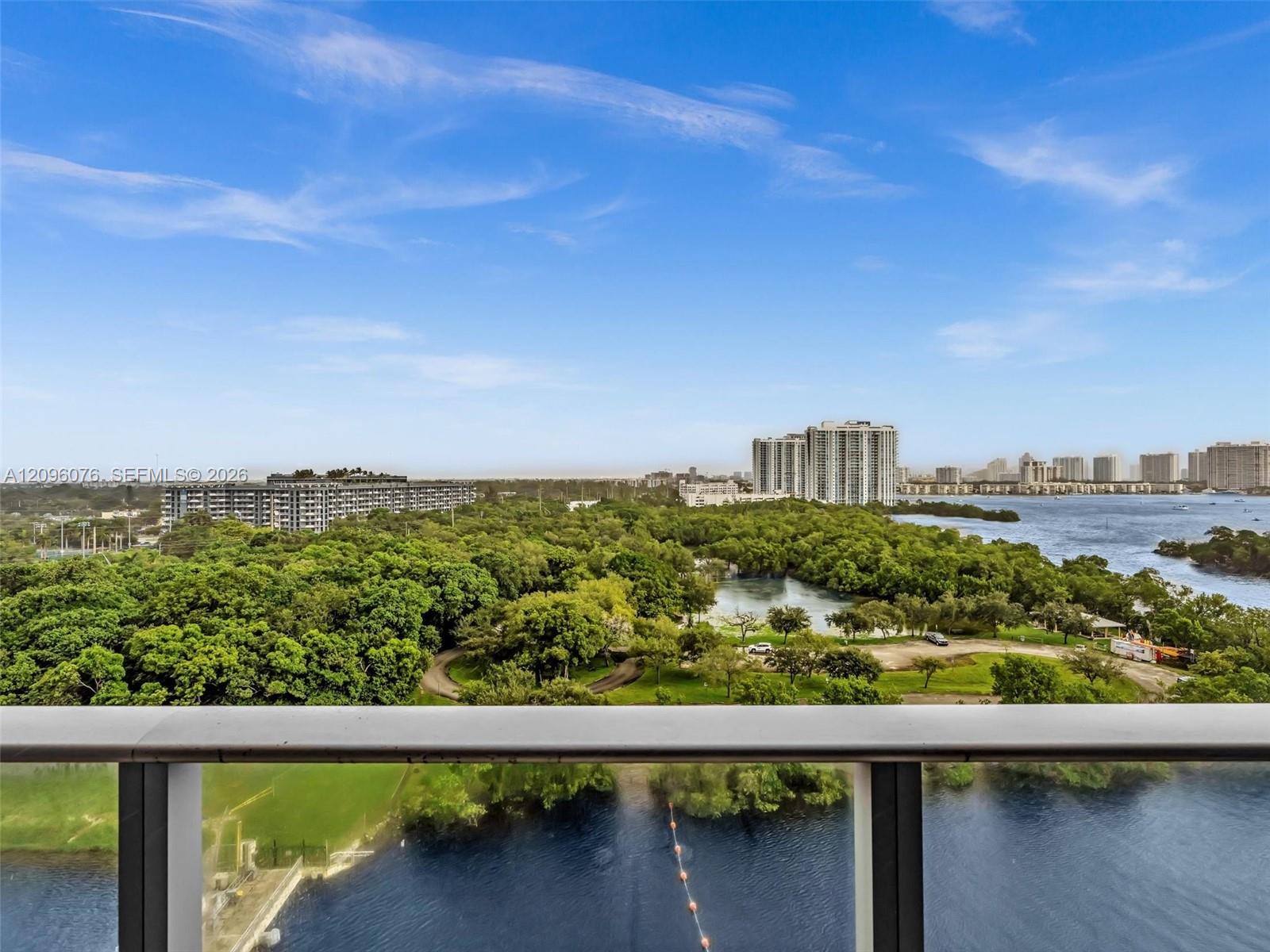 Balcony featuring a glass and metal railing, overlooking a water expanse, expansive green space, and metropolitan skyline