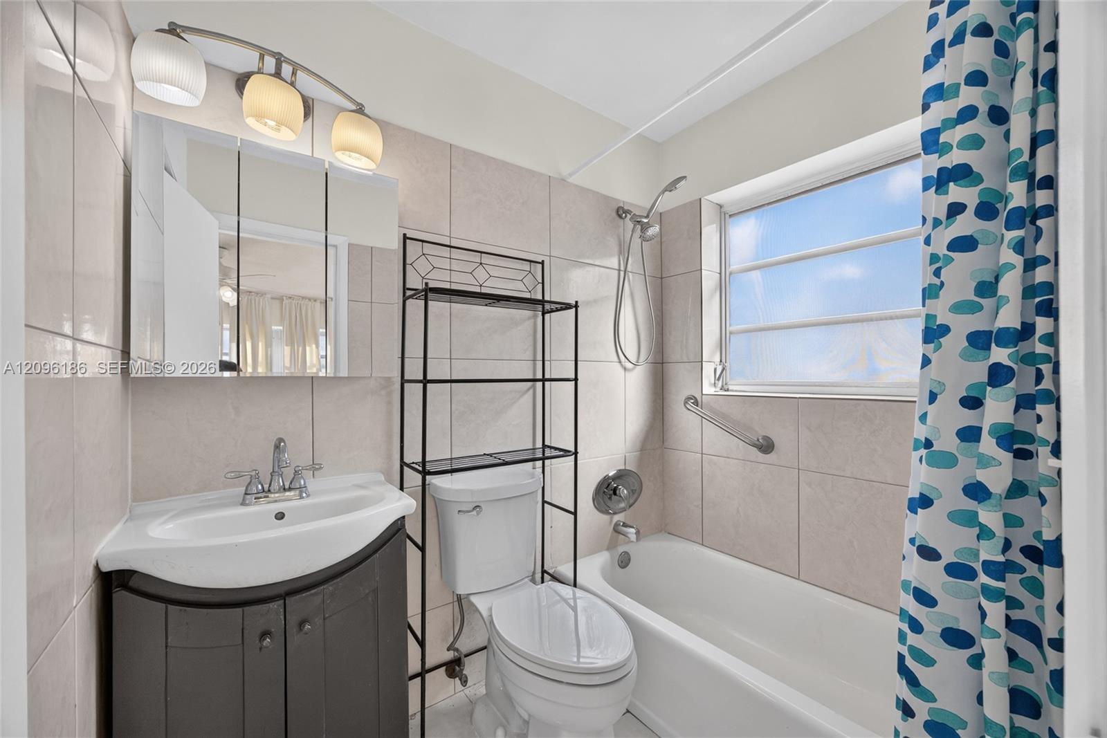 Bathroom featuring a pedestal-style vanity with storage, tile-surround bathtub with a handheld shower head, wall-mounted light fixture, and horizontal-slat window