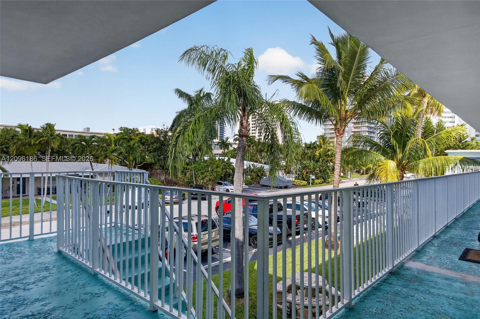 Elevated balcony featuring a protective metal railing, textured flooring, and surrounding mature palm trees