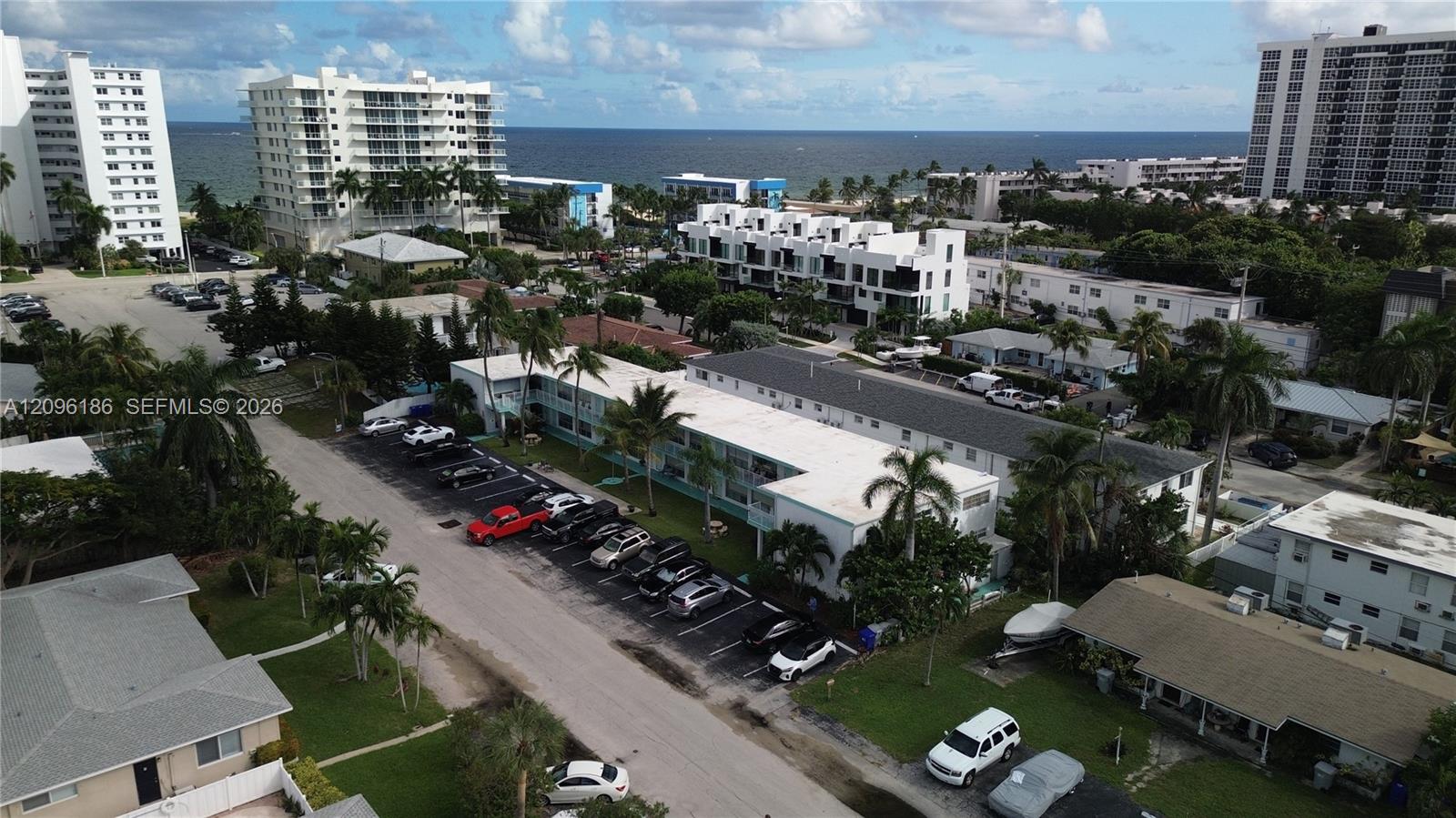 Aerial perspective of a multi-unit residential complex featuring on-site surface parking, mature palm trees, and proximity to the coastline