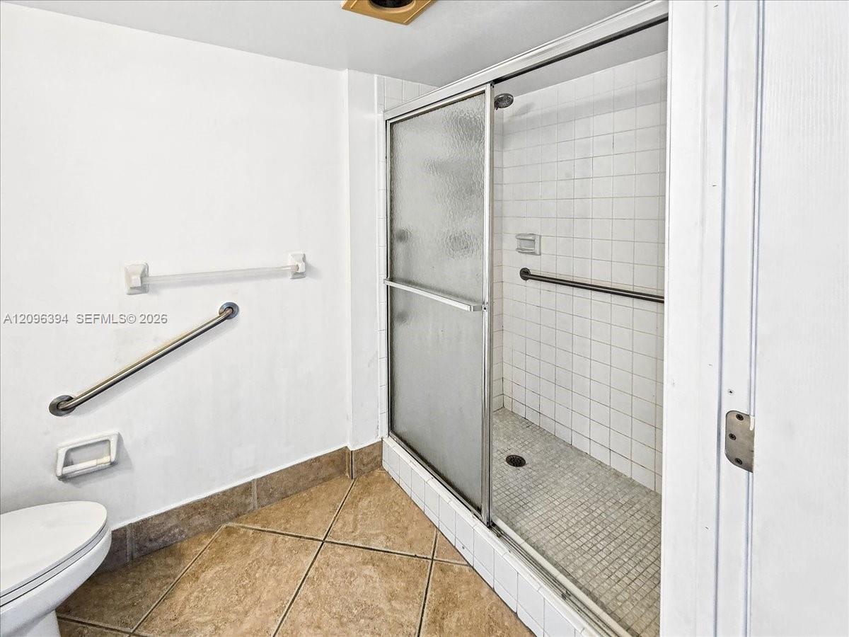 Walk-in shower featuring a sliding glass door, tiled wall surround, and metal grab bars, paired with large-format ceramic floor tiling