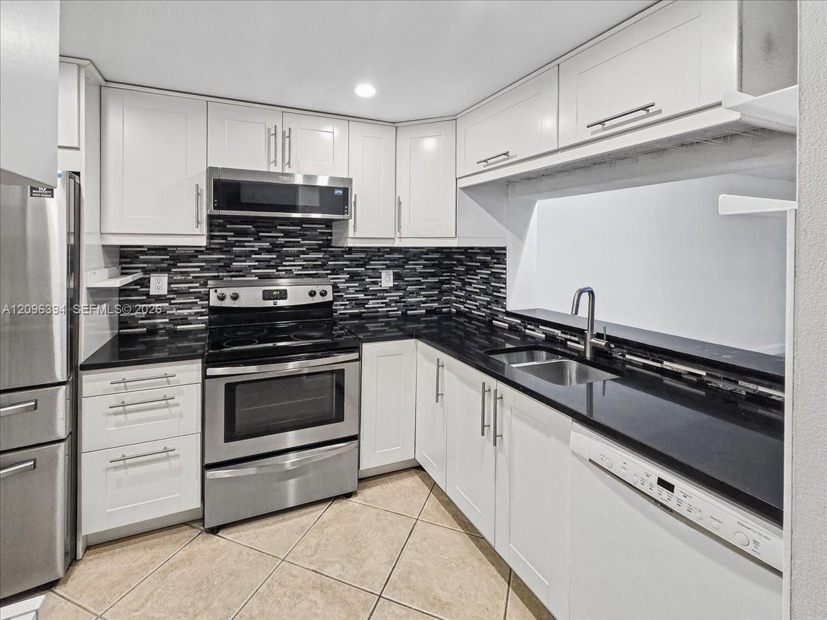 Kitchen featuring white shaker-style cabinetry, black solid-surface countertops, stainless steel appliances, a multi-tonal mosaic tile backsplash, and tile flooring