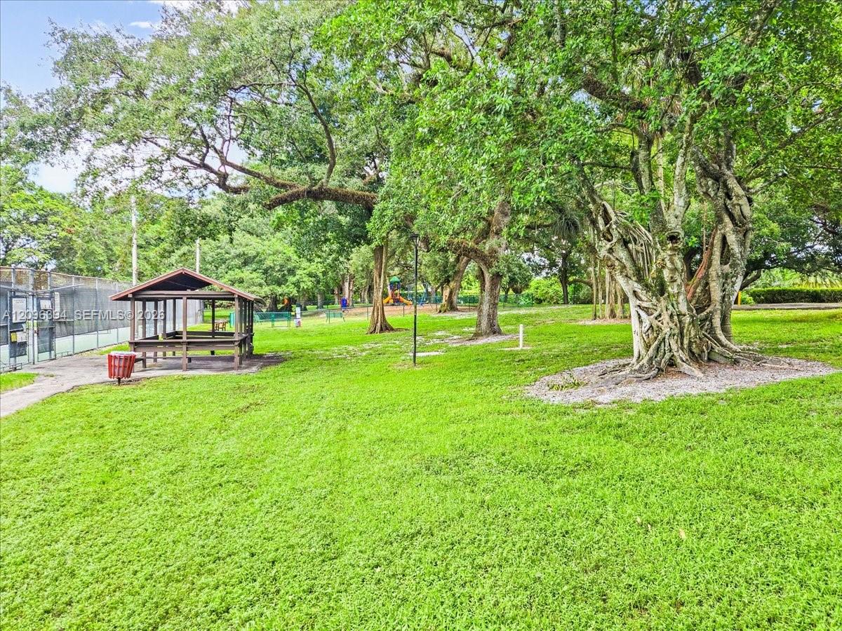 Expansive green space featuring mature canopy trees, a covered wooden pavilion, and a fenced recreation court