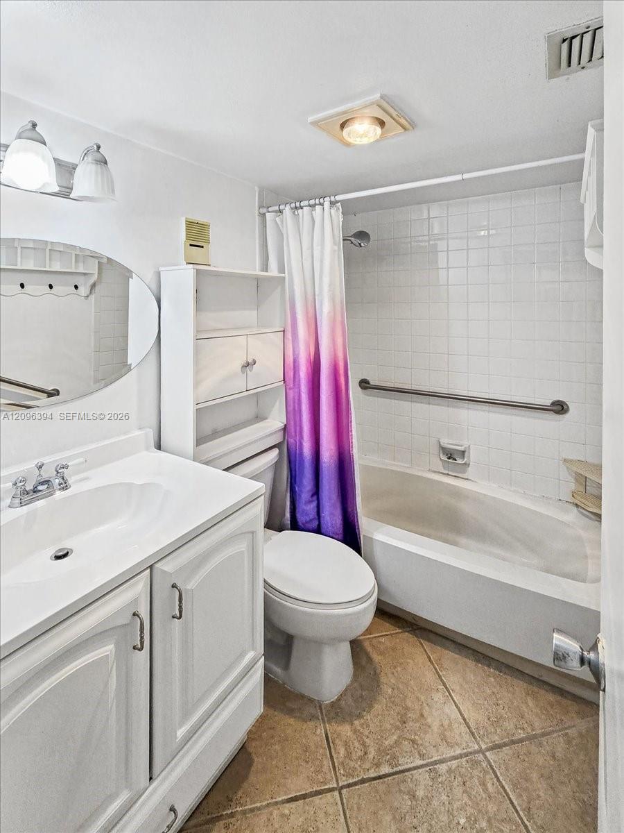 Bathroom featuring a white vanity cabinet, dual-handle faucet, oval wall mirror, tile-surround bathtub with a metal grab bar, and neutral-toned tile flooring