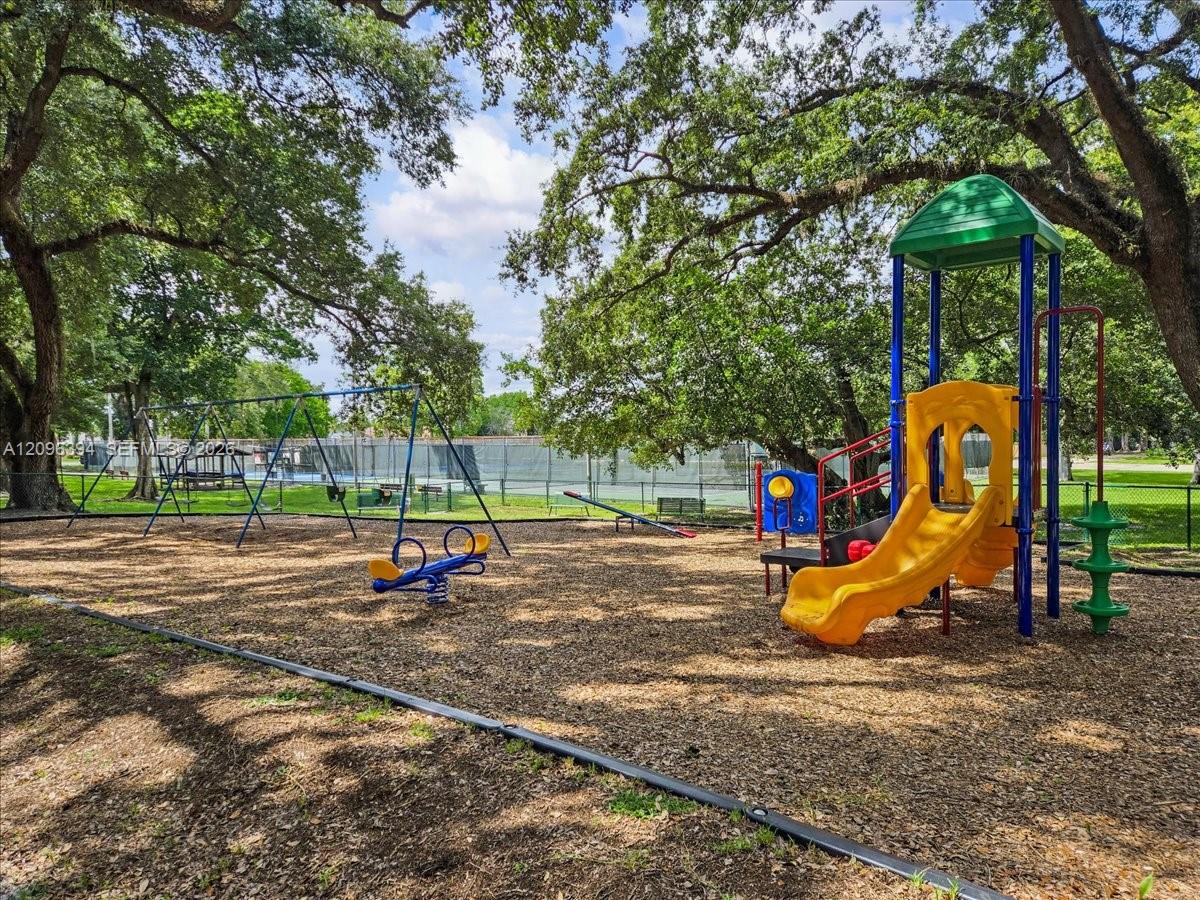 Outdoor recreation area featuring a playground structure with a slide, swings, and a teeter-totter, set within a park-like space with mature trees and perimeter fencing