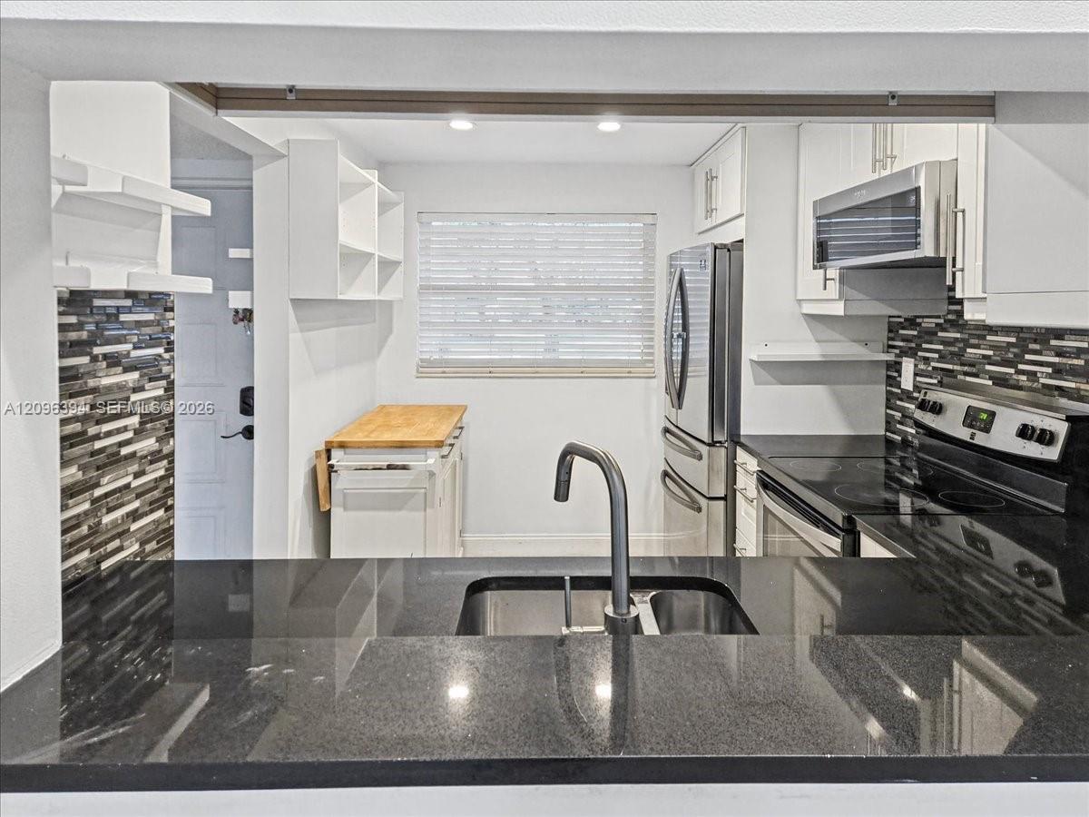 Kitchen featuring black stone countertops, a stainless steel dual-basin undermount sink with a high-arch faucet, white cabinetry, a multi-tone horizontal tile backsplash, and stainless steel appliances