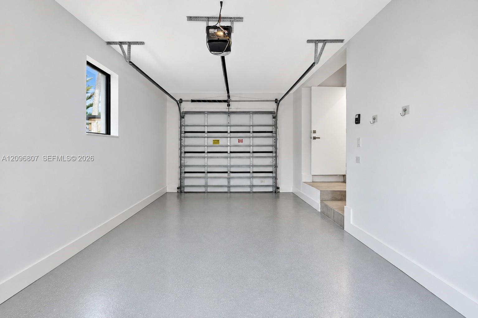 Interior garage featuring smooth white walls, a finished epoxy floor, an automatic overhead door opener, a small rectangular side window, and an interior access door with steps