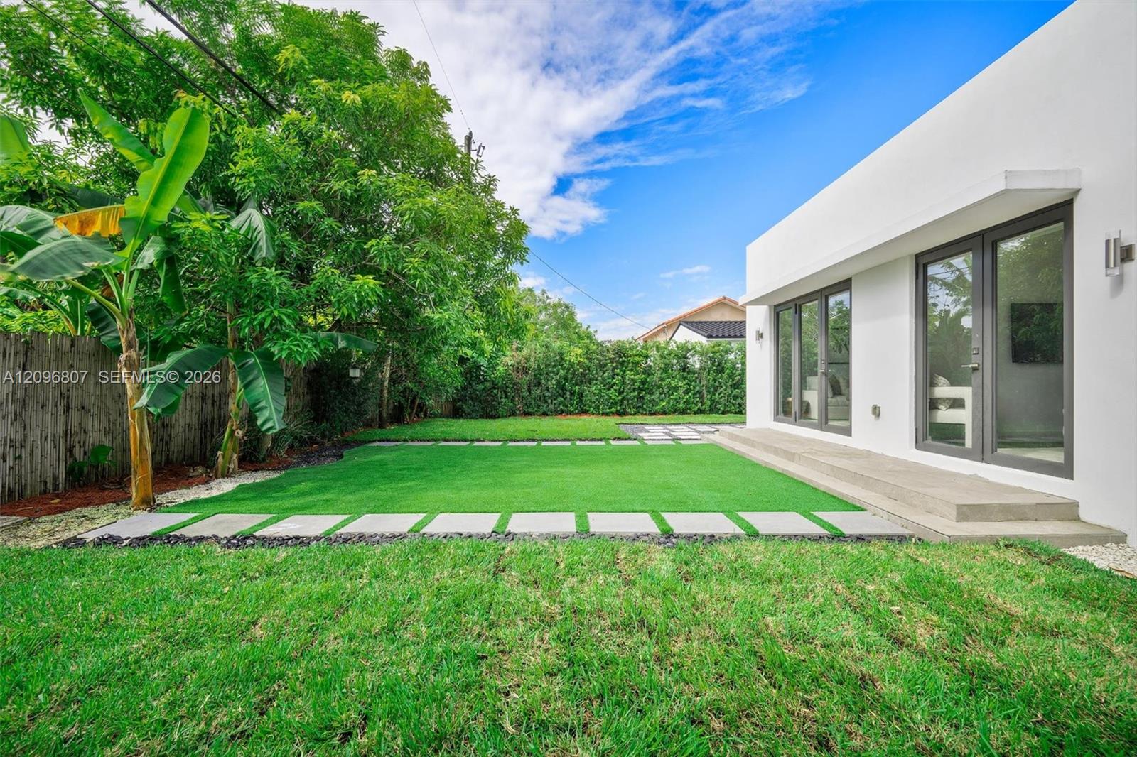 Exterior space featuring a concrete patio, synthetic turf lawn, stone paver pathway, and dual glass-paneled double doors