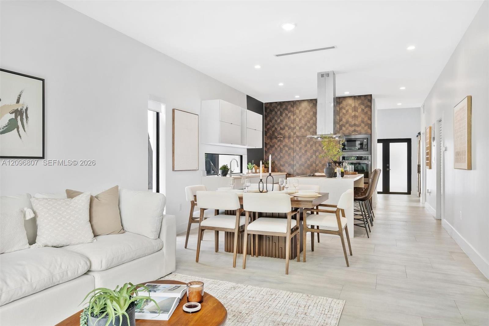 Open-concept kitchen featuring chevron-patterned backsplash tile, center island with stone countertop, stainless steel range hood, built-in wall oven, and light-toned wood-finish flooring