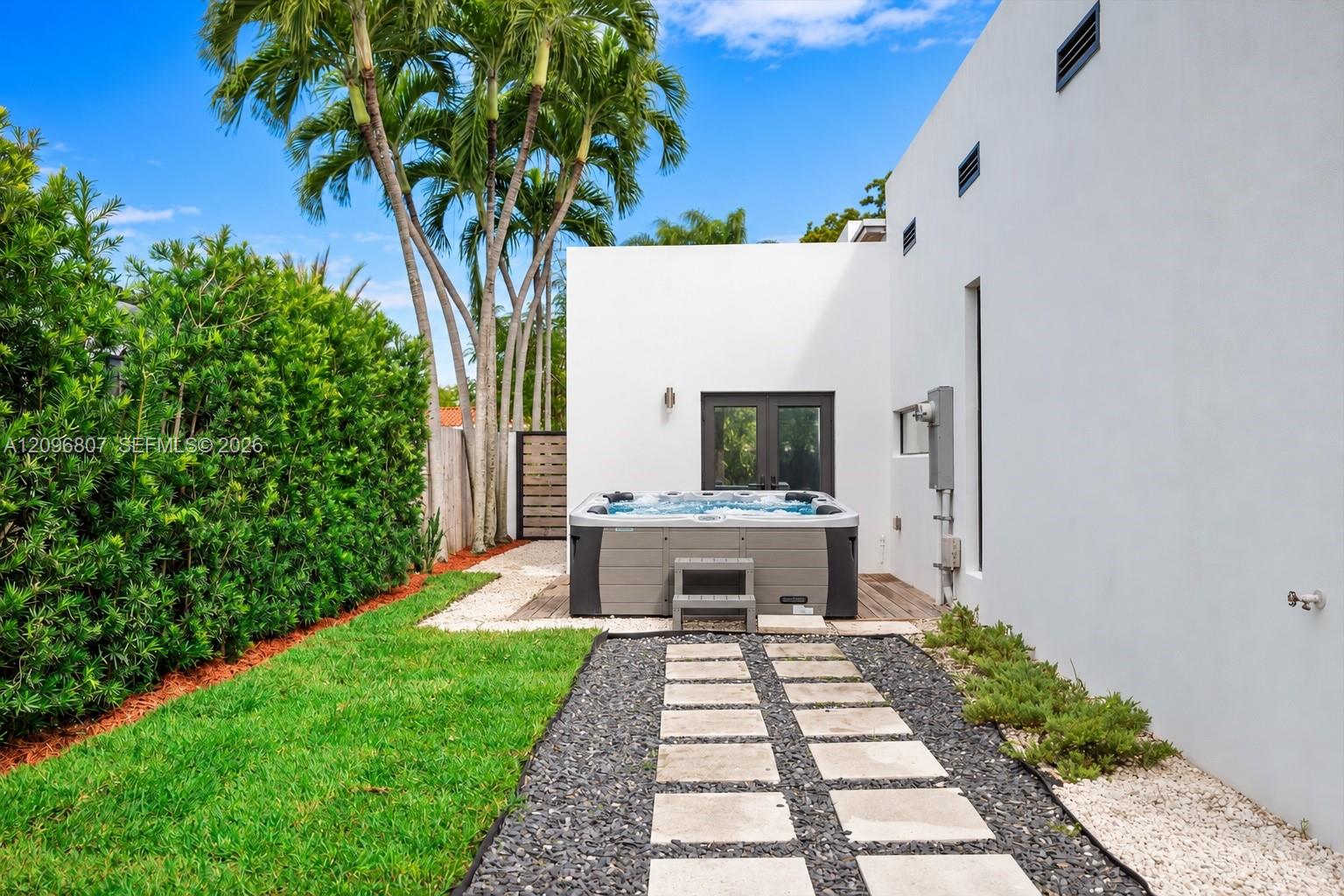 Outdoor space featuring a standalone spa, stone paver walkway with gravel surround, manicured lawn, and mature palm trees Outdoor space featuring a standalone spa, stone paver walkway with gravel surround, manicured lawn, and mature palm trees