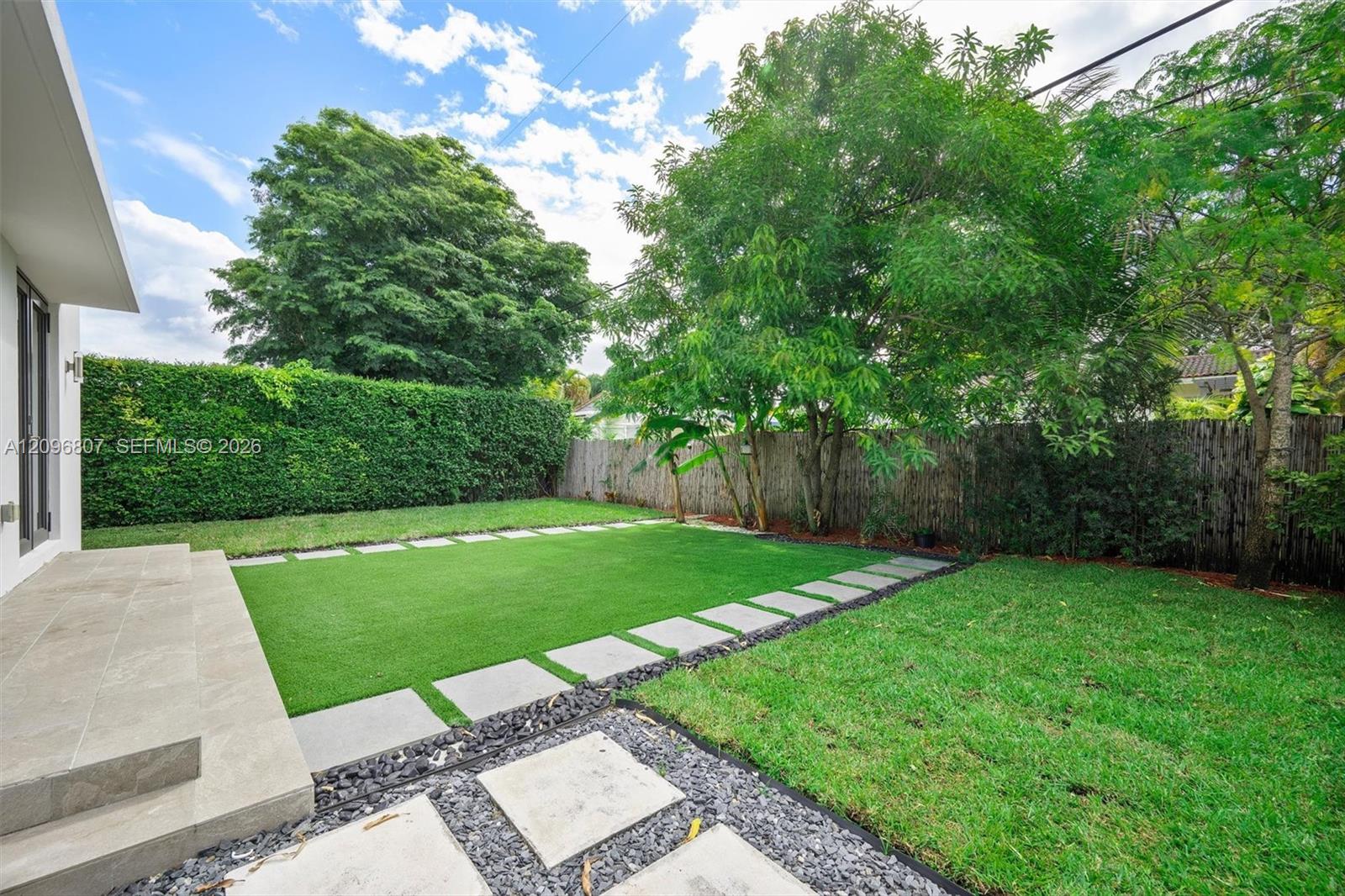 Rear yard featuring a tiled patio, synthetic turf lawn, stone paver pathway, perimeter hedge, and mature trees