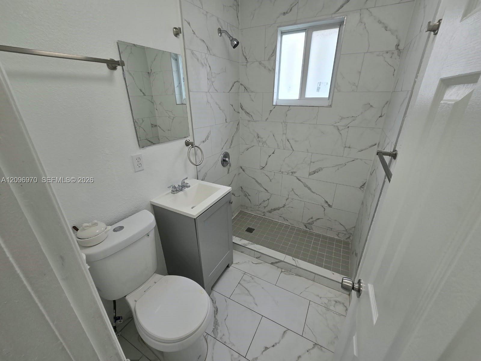 Bathroom featuring floor-to-ceiling large-format tile with veining, grey vanity cabinet, white sink basin, and glass-paneled walk-in shower